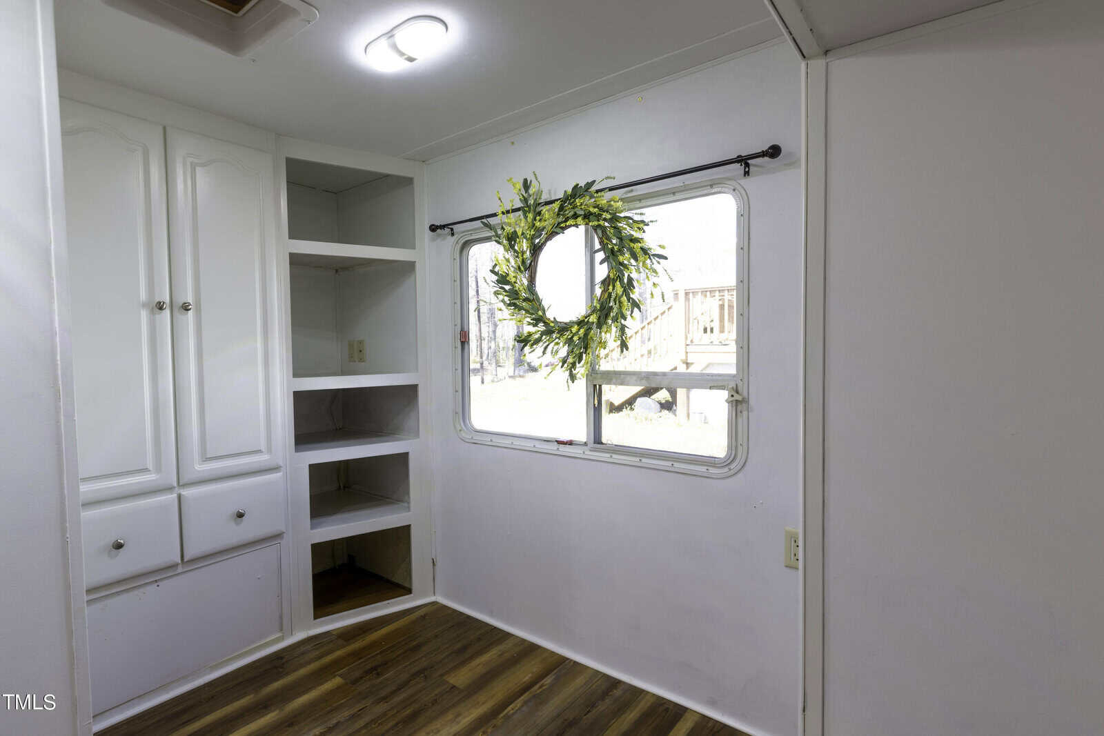 A bright, white room with wood flooring, built-in cabinets and shelves on the left, and a large window decorated with a green and yellow wreath on the right wall.