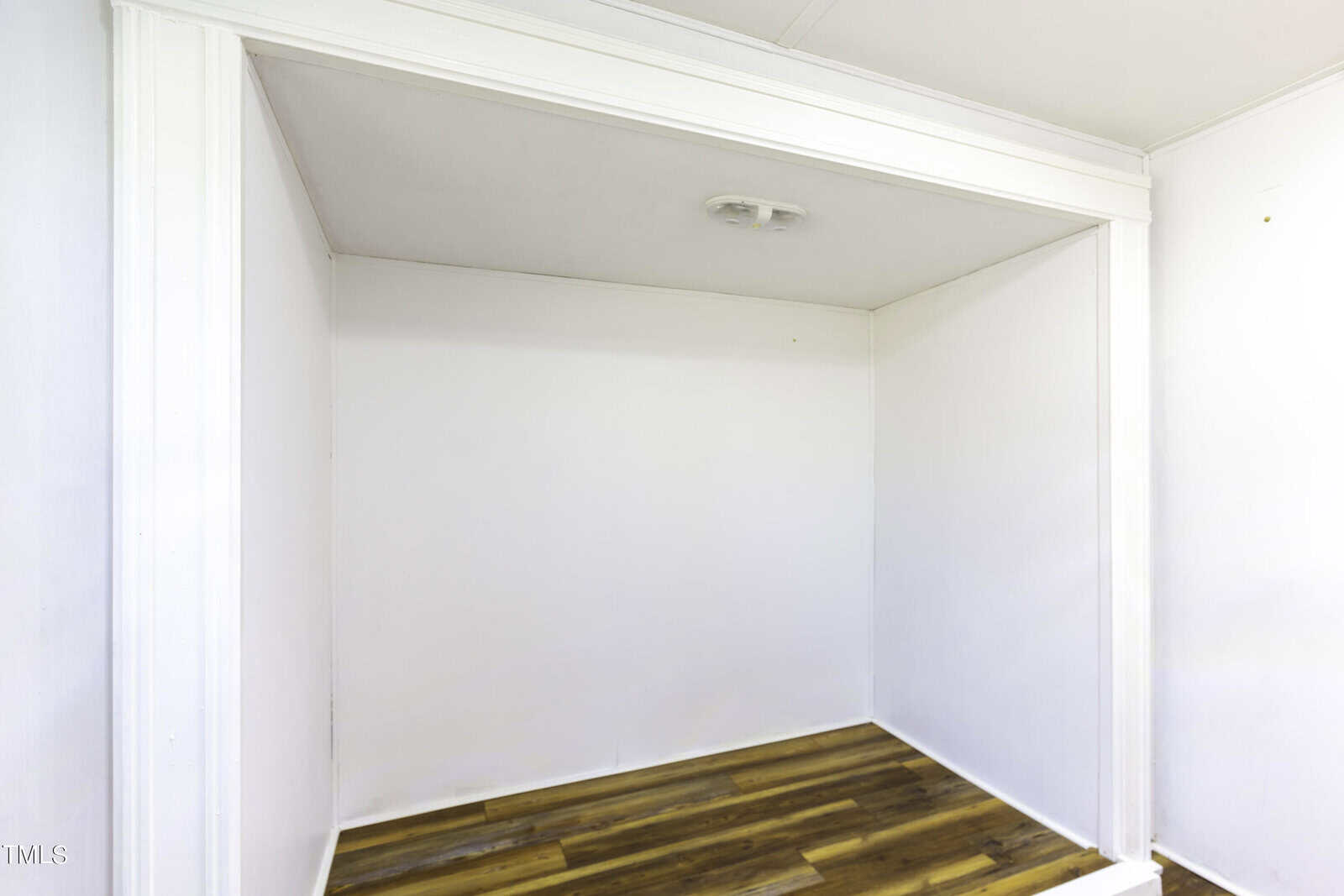 A small, empty white room with clean walls, a low ceiling, and wood-patterned flooring. The room is well-lit with a ceiling light fixture at the center.