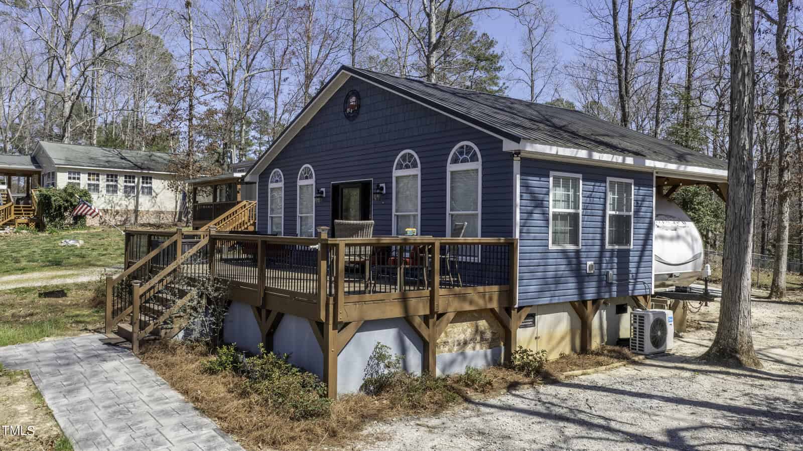 Blue cottage with white trim and arched windows, elevated on a wooden deck with stairs. There are trees around, a small pathway, and another house with an American flag in the background on the left.