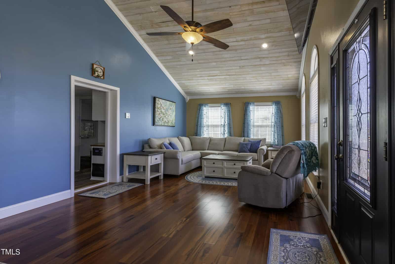 A cozy living room with teal accent walls, wood ceiling, dark wood floors, a ceiling fan, a sectional sofa, an armchair, and large windows with teal curtains letting in natural light.