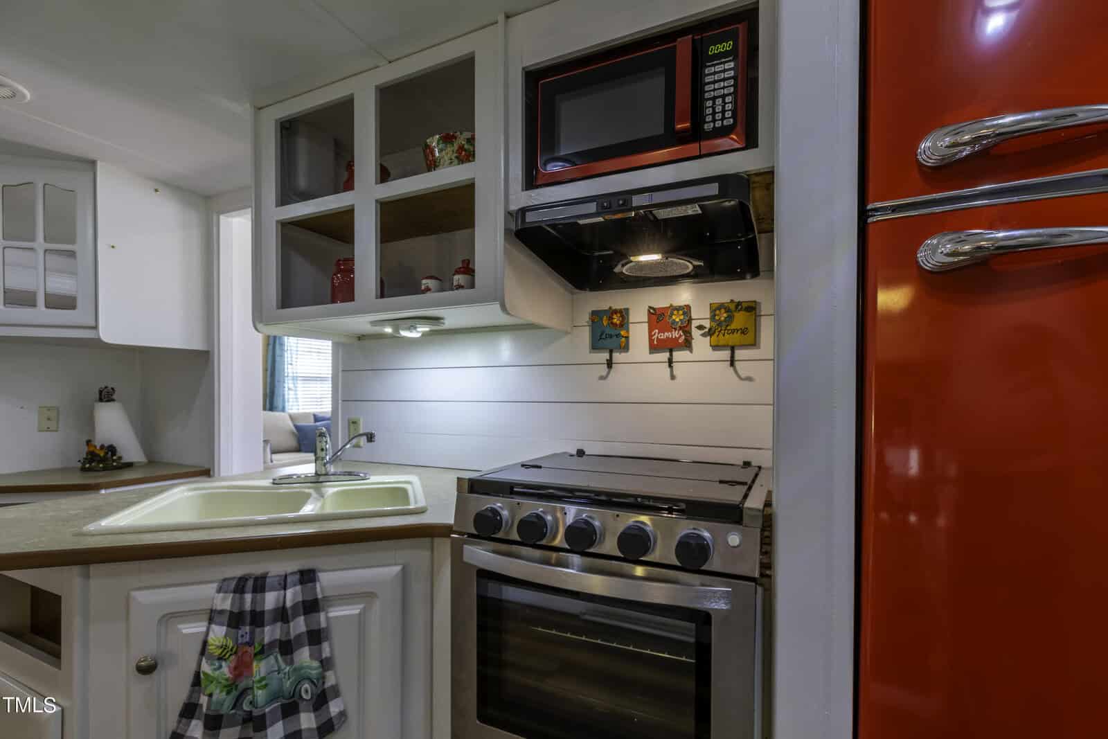 A compact vintage kitchen features a red refrigerator, stovetop oven, microwave, double sink, open shelves with mugs, and colorful decorative hooks hanging above the stove. A dish towel hangs on the cabinet door.