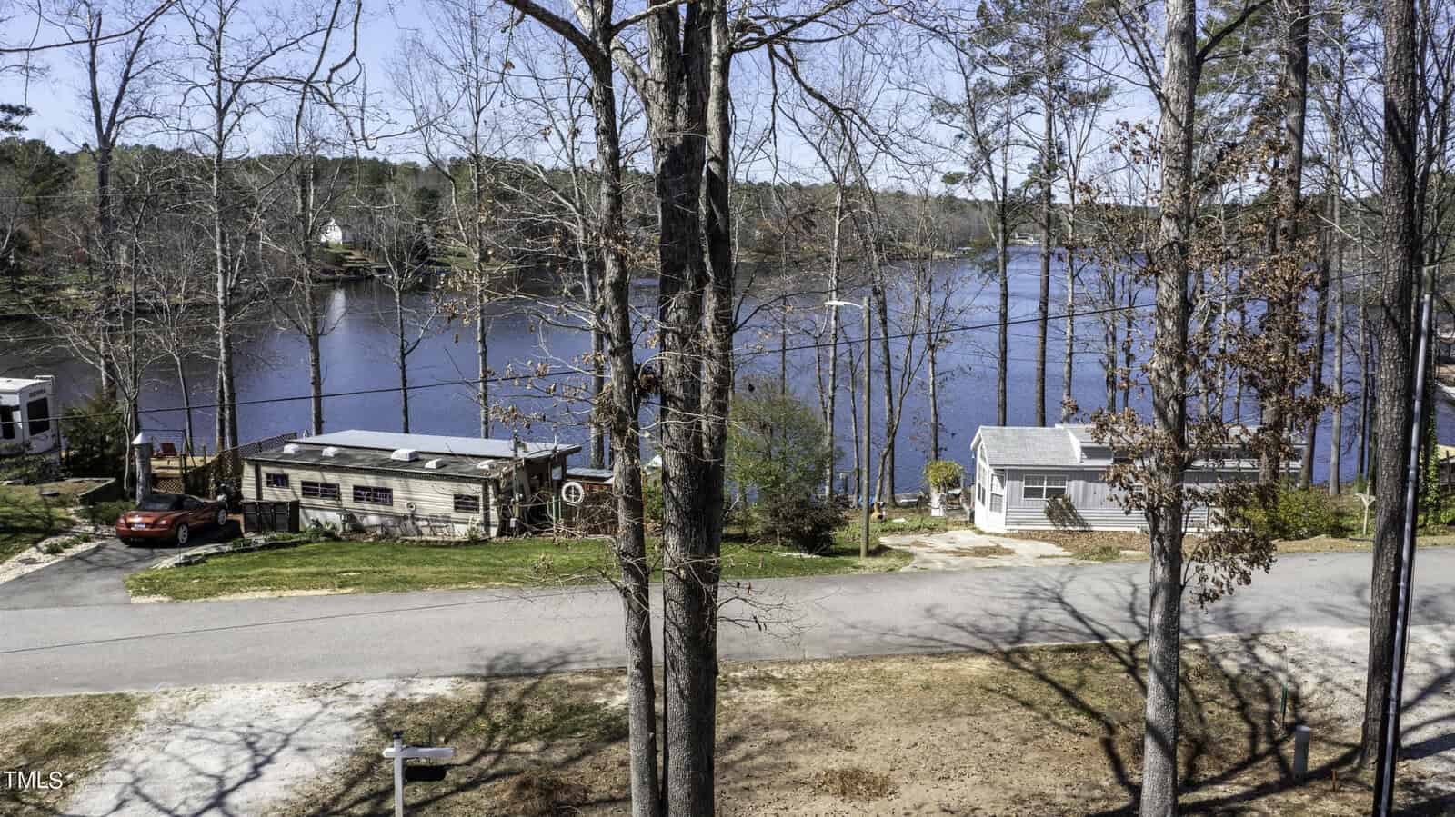 View of two small houses near a lake, with a road in front, leafless trees, a red car parked to the left, and a wooded shoreline in the background under a clear sky.