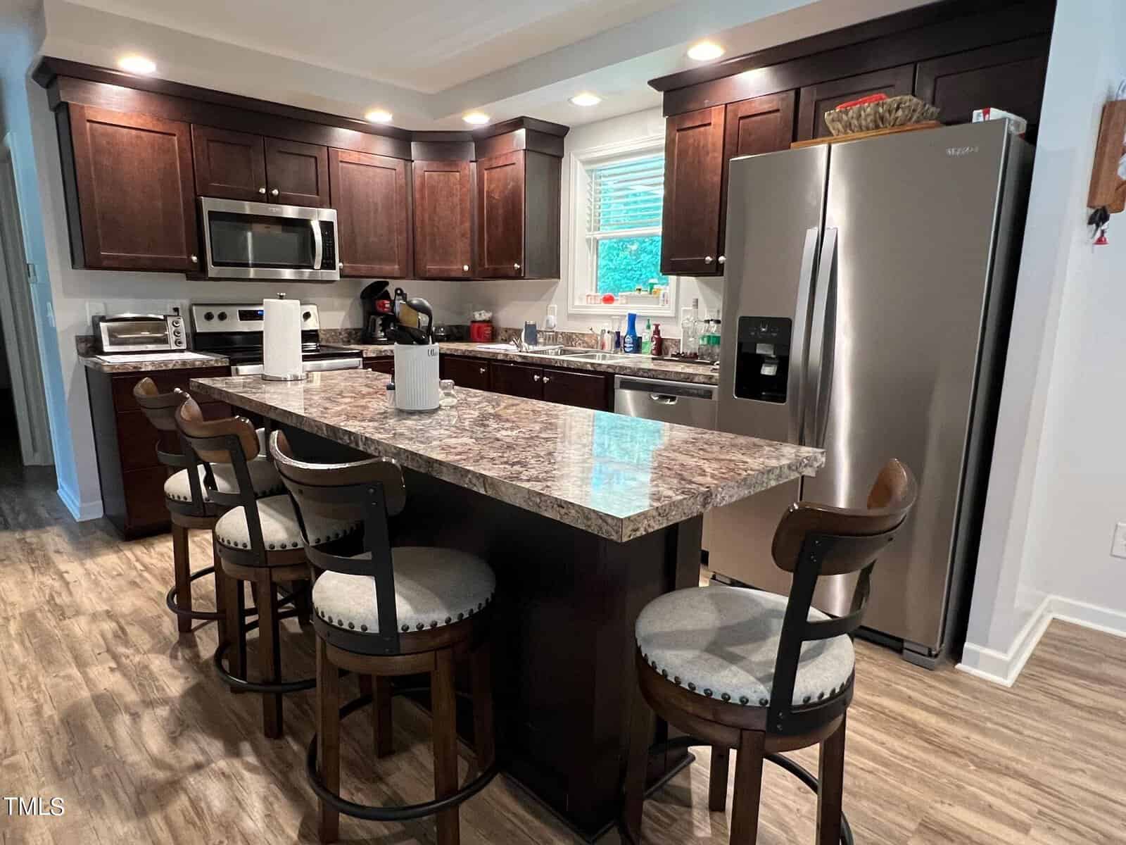 A modern kitchen featuring dark wood cabinets, stainless steel appliances, and a marble countertop island with four cushioned bar stools. The floor is wooden, and the space is well-lit with recessed lighting. The kitchen includes a microwave, oven, and refrigerator.