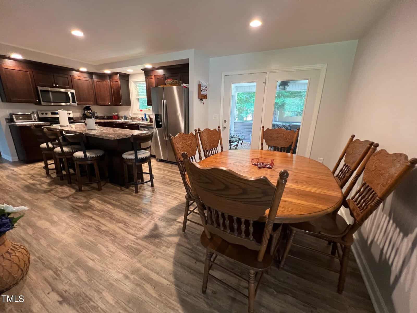 A modern kitchen with dark wood cabinets, stainless steel appliances, and a granite island with barstools. Adjacent is a dining area with a wooden table and six matching chairs. French doors open to an outdoor space, and the floor is covered with light wood.