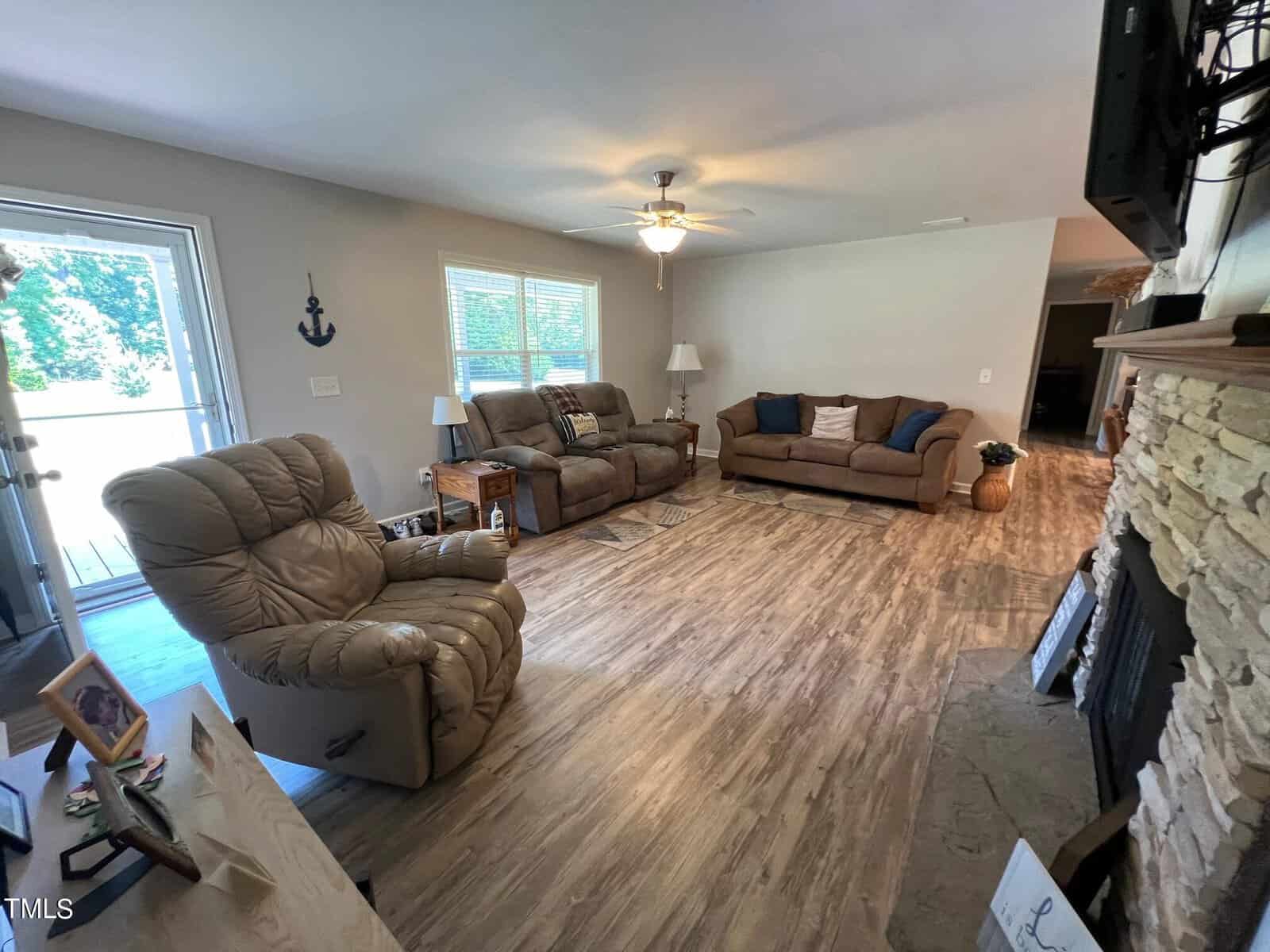 A cozy living room with wooden flooring, featuring two brown sofas with assorted pillows and a recliner next to a window with an anchor decoration. A large TV is mounted above a stone fireplace. There is a door leading outside, and various decor items are placed around the room.