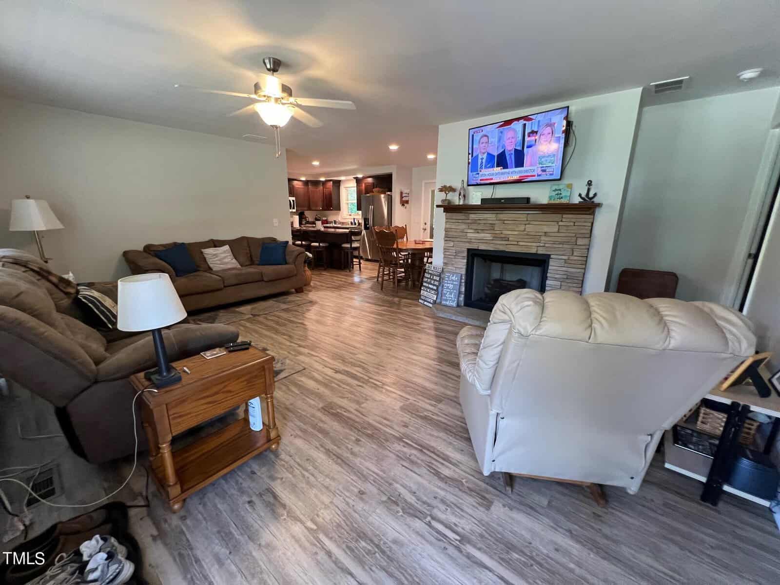 A spacious living room featuring a ceiling fan, two recliners, a sofa, and a lamp on a wooden side table. The floor has wood-like tiles. A fireplace with a mounted TV is on the right wall. The background shows a dining area and a kitchen with dark cabinets.