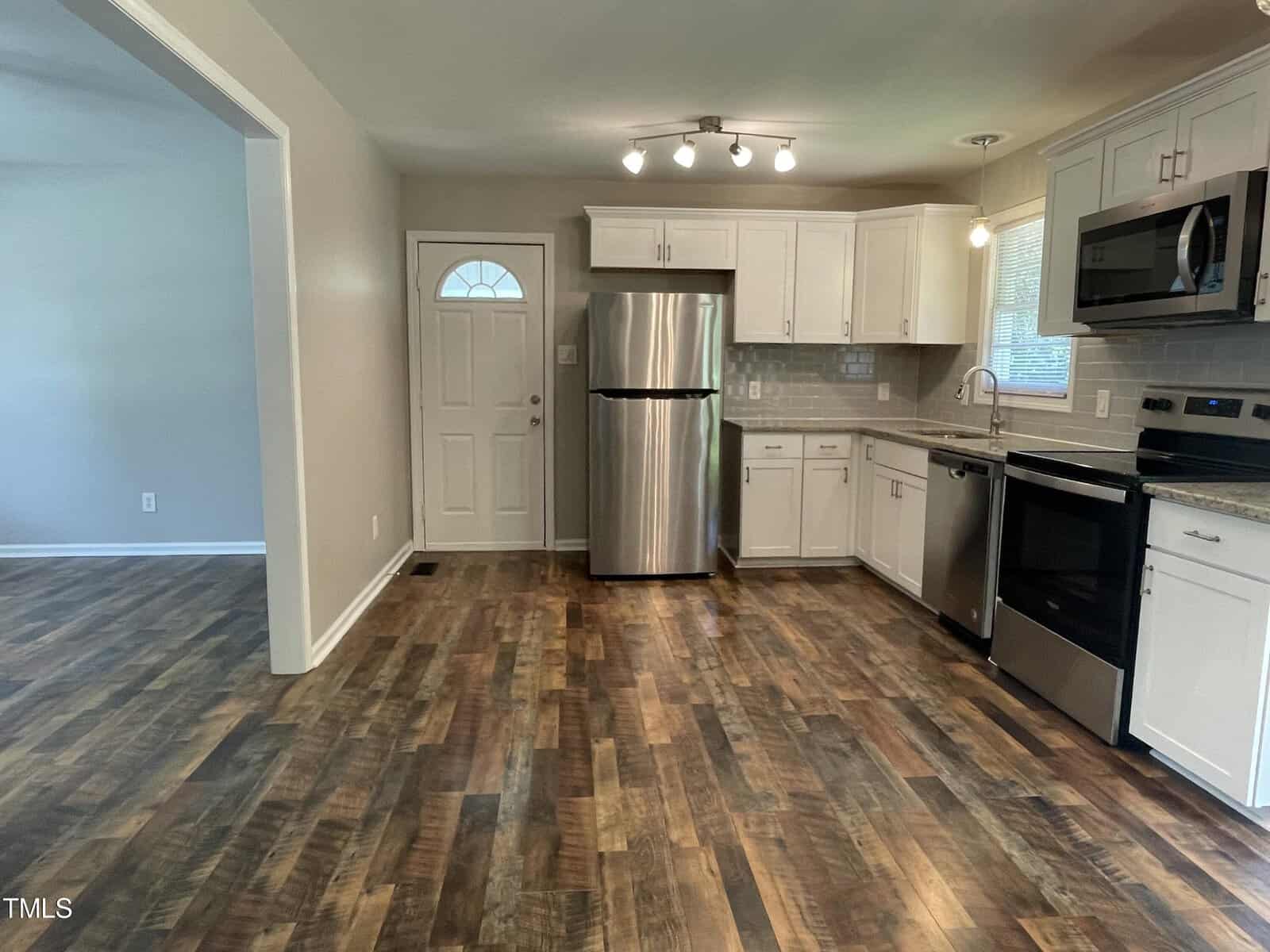 A modern kitchen with stainless steel appliances, including a refrigerator and oven. White cabinets, granite countertops, and light wood flooring are visible. A door and window let in natural light, with recessed ceiling lights above.