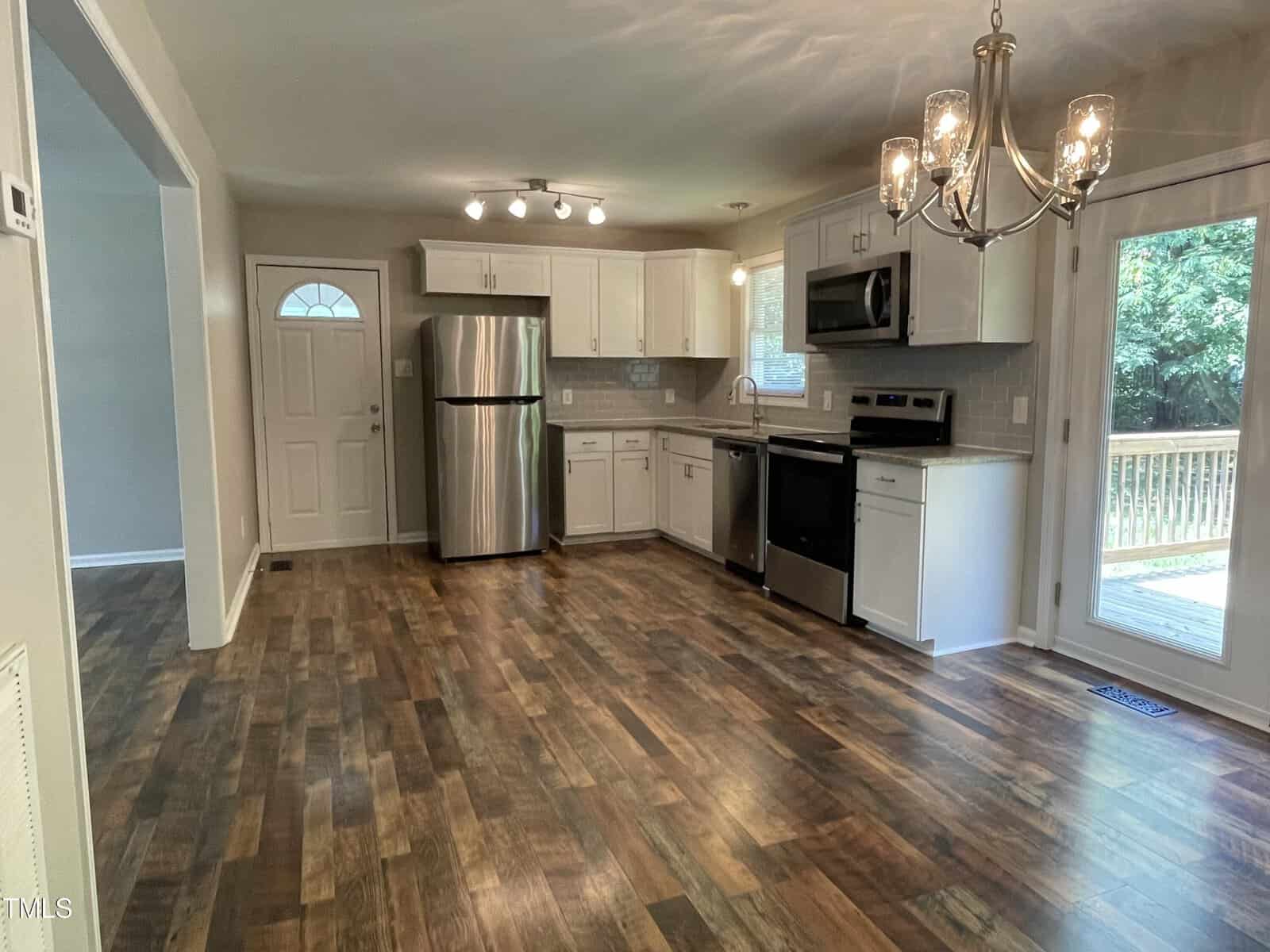 Modern kitchen with wooden flooring, stainless steel appliances, white cabinets, and a tile backsplash. A chandelier hangs over the space, with a door leading to a deck and trees visible outside.