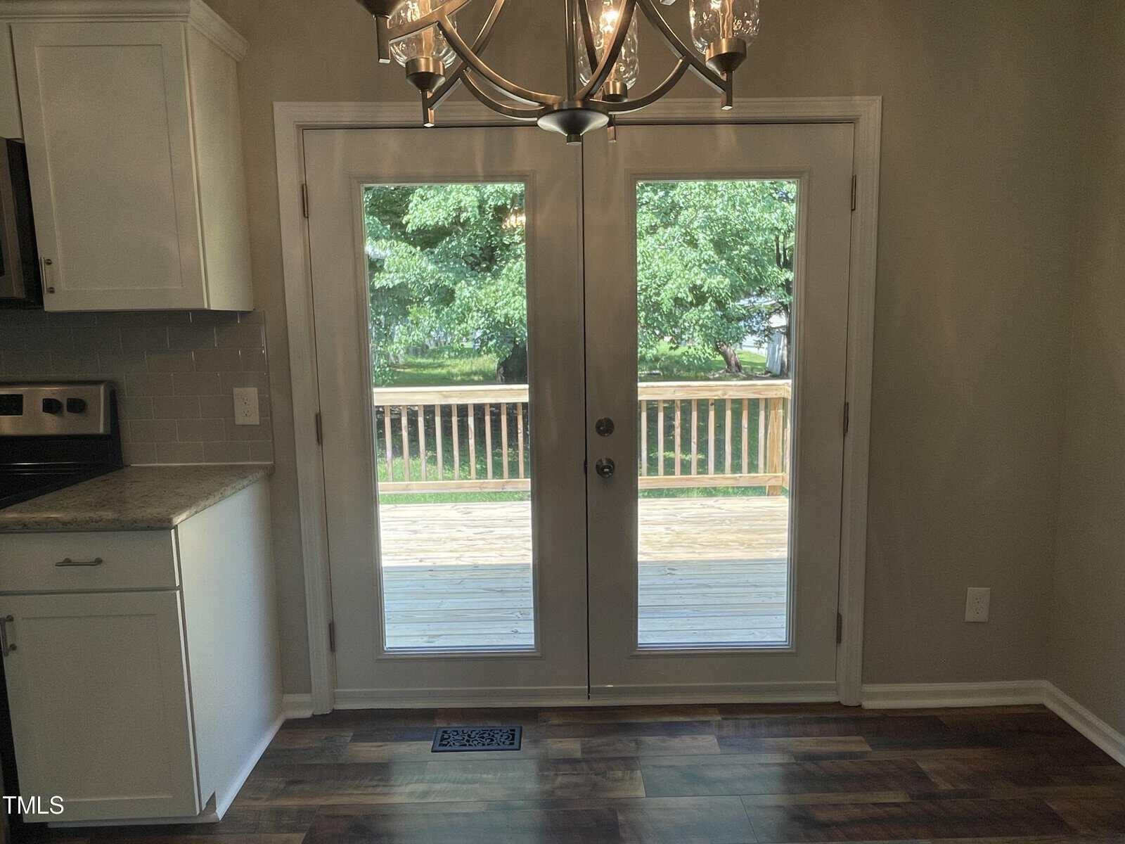 A kitchen area with white cabinets and a tiled backsplash, featuring double glass doors leading to a wooden deck outside. A chandelier hangs above, and through the doors, green trees and a yard are visible.
