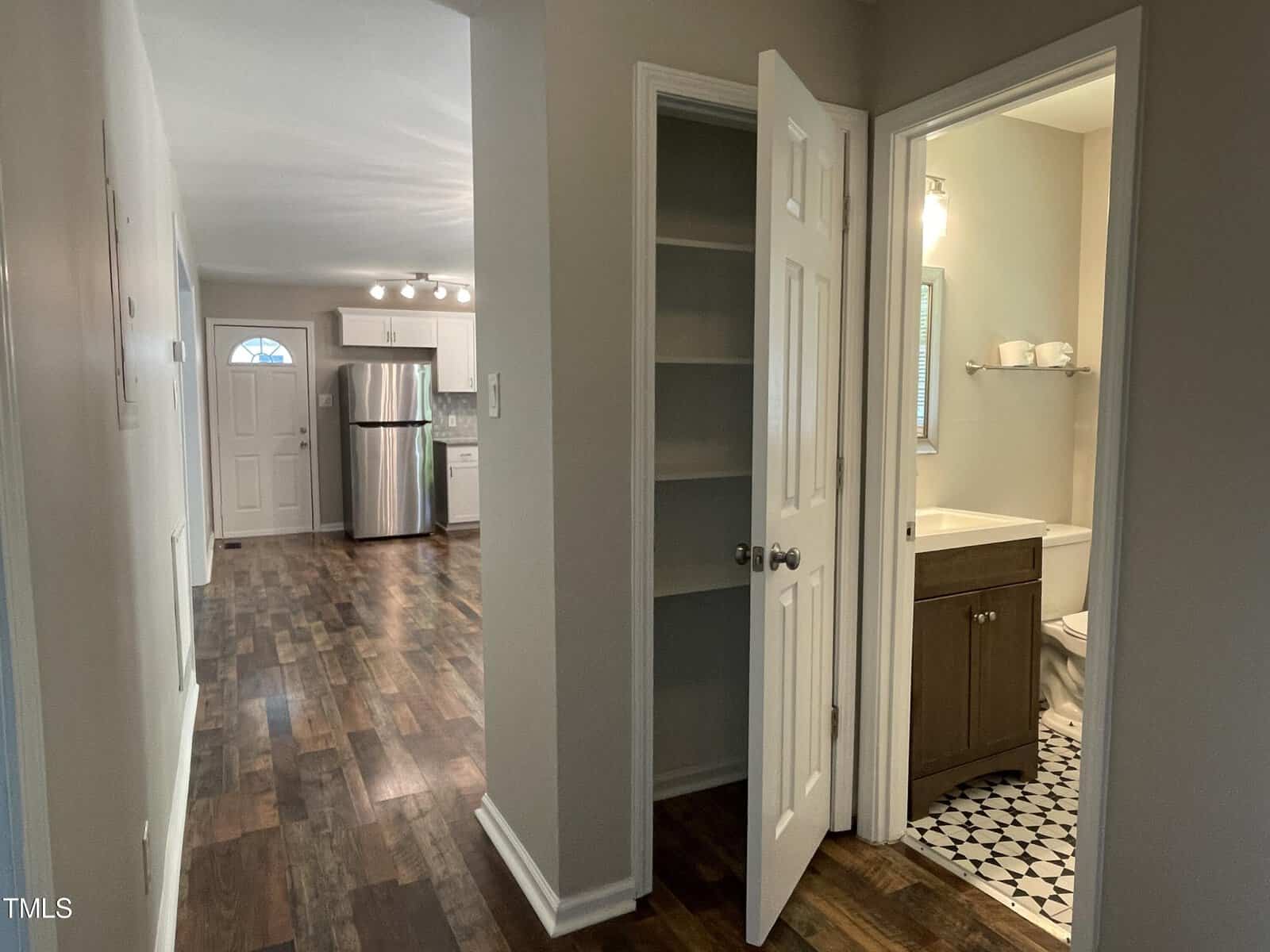 A hallway with a wooden floor leading to a kitchen with stainless steel appliances. An open pantry door is in the foreground, and a bathroom with a patterned floor is visible to the right.