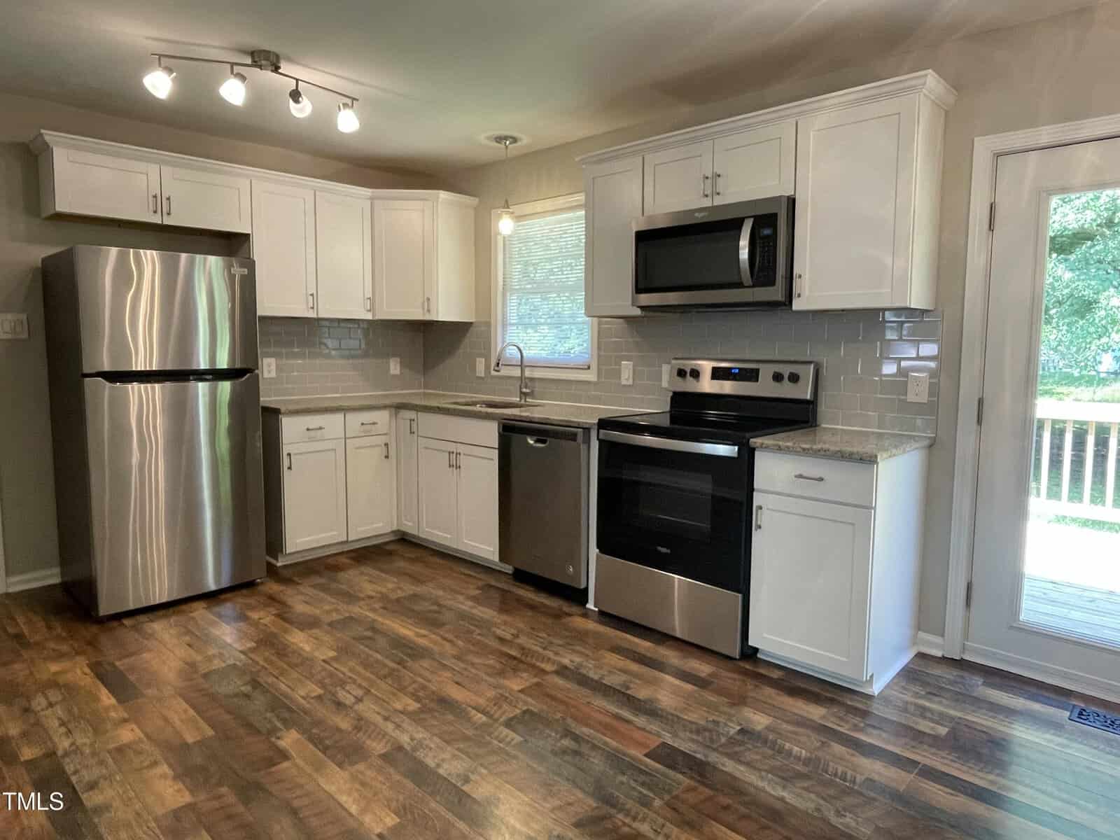 A modern kitchen featuring white cabinets, stainless steel appliances, including a refrigerator, dishwasher, oven, and microwave, with a gray tile backsplash and hardwood floors. A glass door on the right leads to an outdoor area.