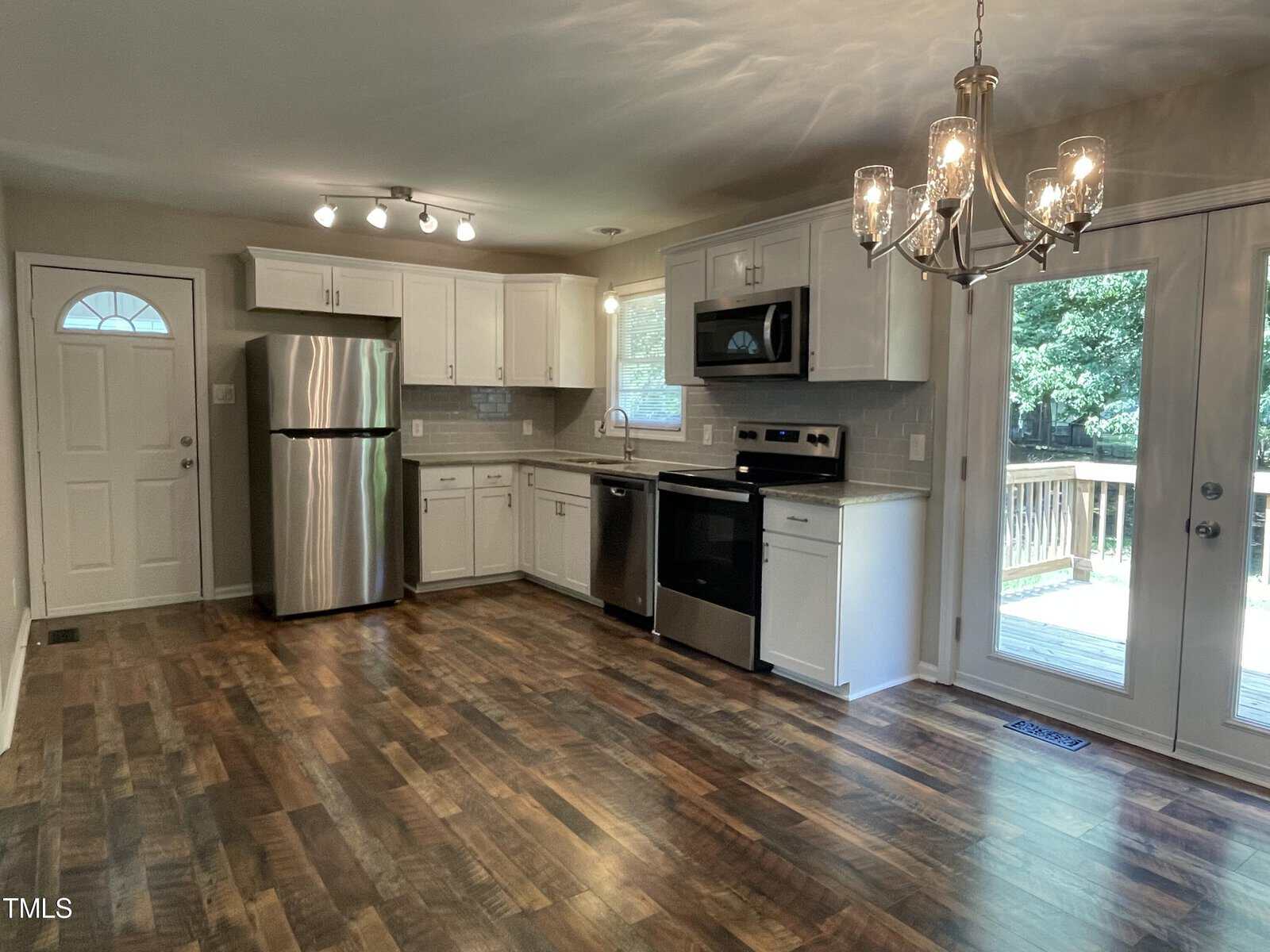 A modern kitchen with stainless steel appliances, including a fridge, oven, and microwave. White cabinets and granite countertops complement the wood-patterned floor. A chandelier hangs above, and glass doors lead to a deck outside.