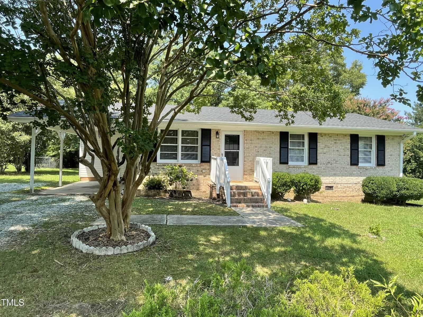 A light brick, single-story house with black shutters sits at 115 Jones Street in Oxford. Bordered by bushes and a large tree, it features steps leading to a central door and a small carport on the left. The grassy yard is complemented by a concrete path, enhancing its charming appeal.