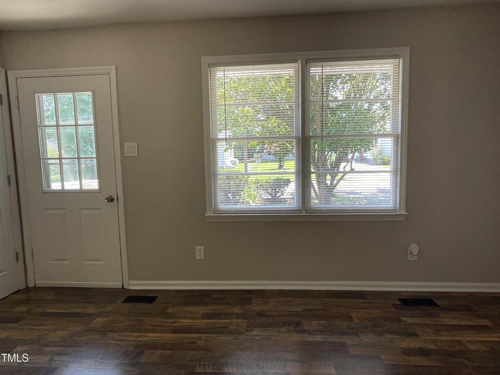 A room with light-colored walls features a door with a window on the upper half and a large, double-paned window. The floor is dark wood, and sunlight illuminates the space through the windows, offering a view of trees and the outdoors.