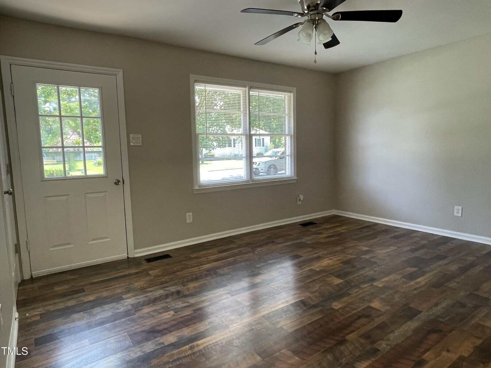A vacant room with wooden floors, light gray walls, and a ceiling fan. A window and a door provide natural light, overlooking greenery outside. The space has a clean and simple appearance.