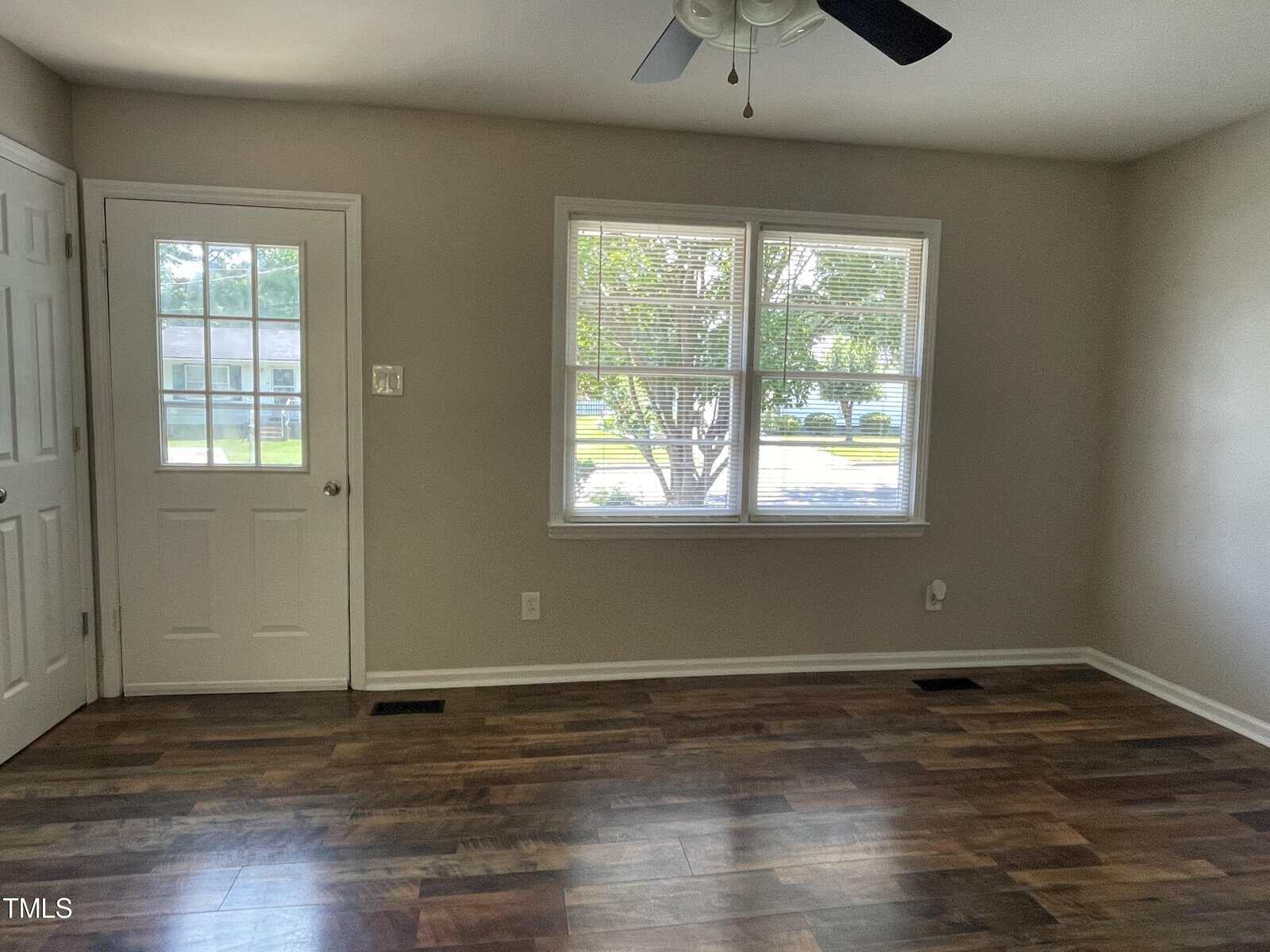 A bright room with wooden flooring, a ceiling fan, and a window showing a view of trees and a road. There is a closed white door on the left and light gray walls. Natural light illuminates the space.
