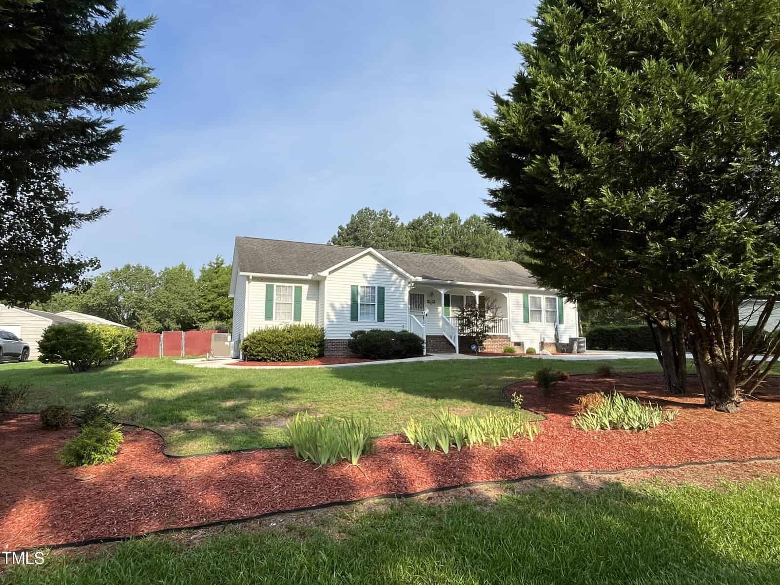 A single-story house with light-colored siding and light blue shutters sits amid a well-maintained lawn with trees and shrubs. Red mulch outlines the garden beds. The sky is clear, indicating sunny weather.