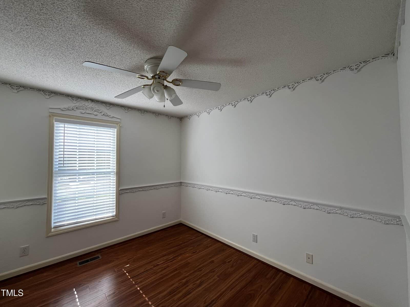 A small, empty room with white walls and decorative molding. It features a single window with blinds, and a ceiling fan with five blades. The floor is hardwood, and natural light streams in, creating a cozy atmosphere.