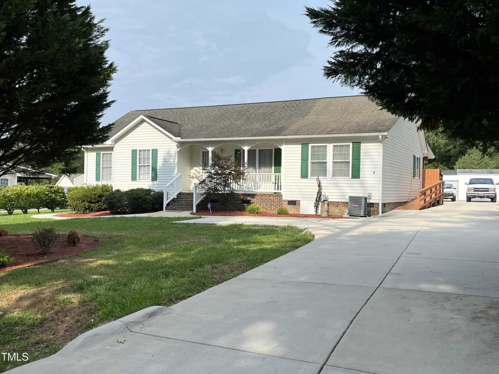 A single-story white house with green shutters and a front porch. It features a large concrete driveway and is surrounded by a landscaped yard with bushes and trees. A car is partially visible in the driveway.