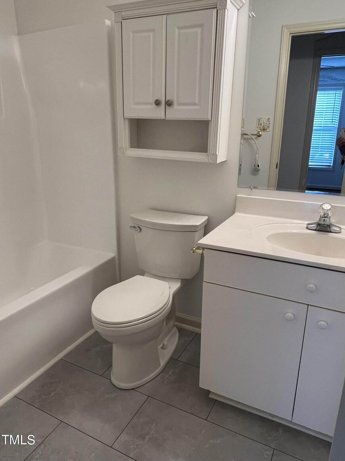 A small, white bathroom features a toilet beside a sink with a cabinet and a chrome faucet. There is a white wall-mounted cabinet above the toilet and a bathtub with a shower curtain rod on the left. The floor is covered with gray tiles.