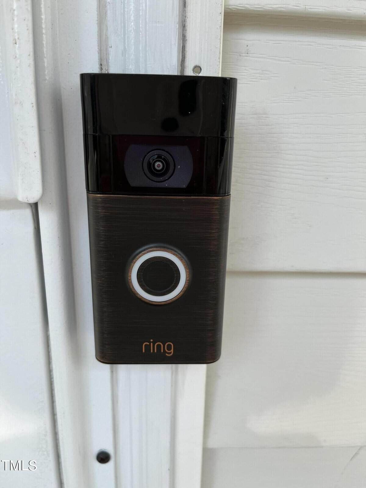 A Ring video doorbell mounted on a white door frame. The device features a camera at the top and a button with an illuminated ring below it.