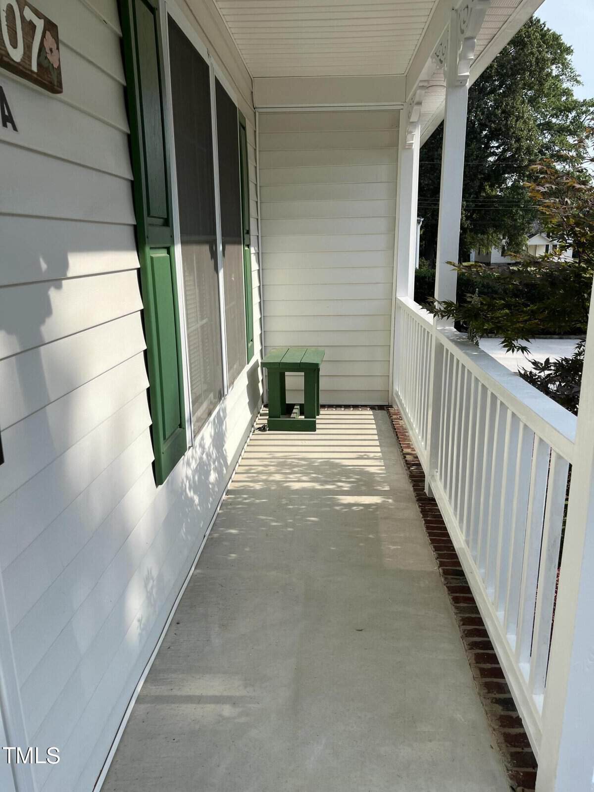 A narrow porch with white siding and a green bench at the end. Green shutters frame the windows on the left, and a white railing encloses the space. Sunlight casts shadows across the concrete floor.