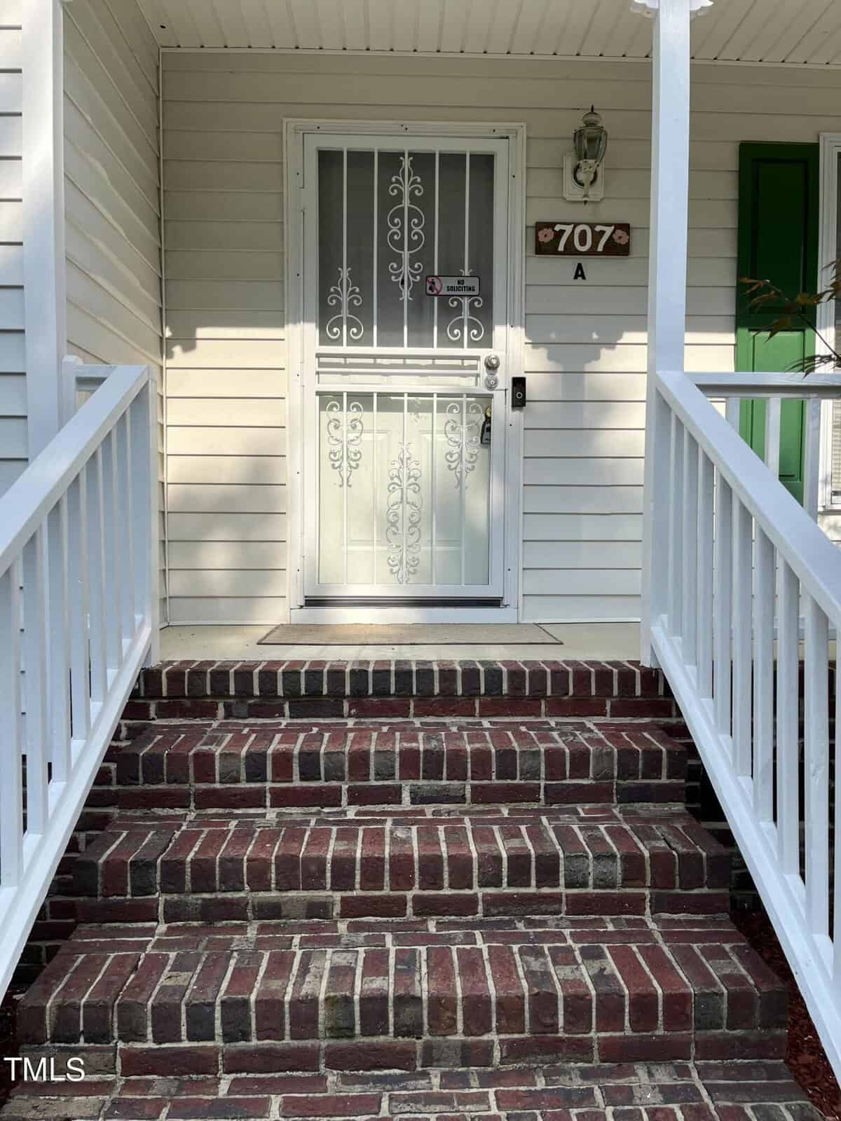 A white door with decorative metalwork is flanked by a small porch area. Brick steps with white railings lead up to the entrance. A sign with "707 A" is visible above the door. The exterior siding is light-colored.