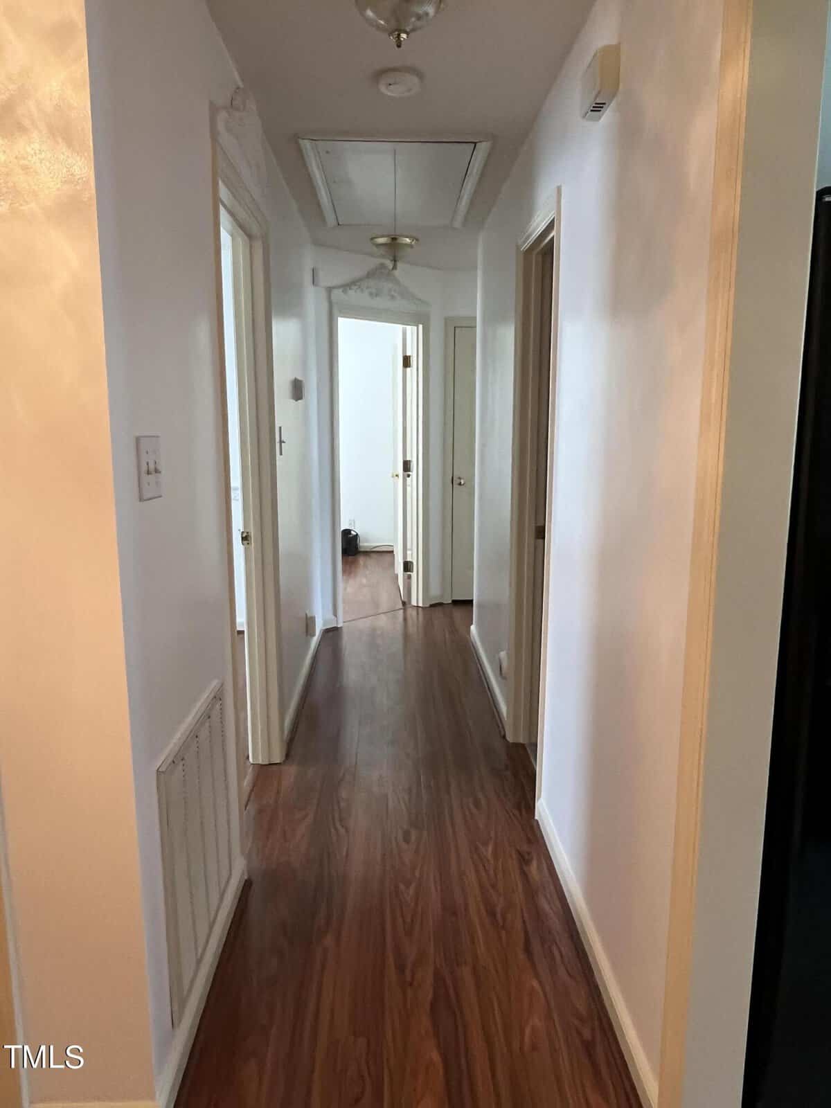 A hallway with wooden flooring and white walls leads to open doors at the end. The ceiling features a white attic hatch and light fixture. There is a vent on the left wall and the hallway receives natural light from the rooms.