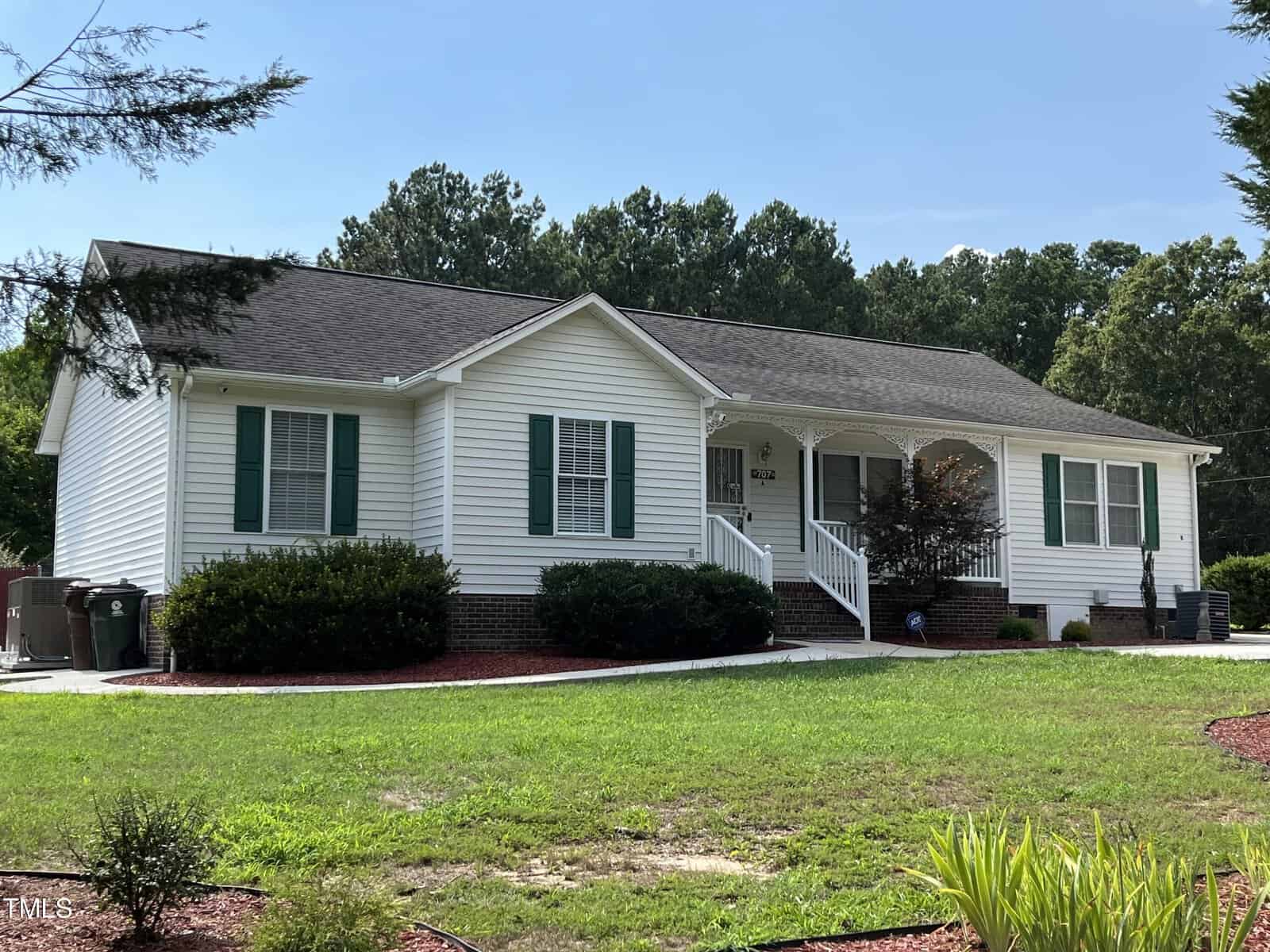 This charming white single-story house at 707 N Pine Street, with its gray roof and quaint front porch, is nestled in Creedmoor. Surrounded by lush green shrubs and a well-maintained lawn, it offers a serene view of trees under a clear blue sky.