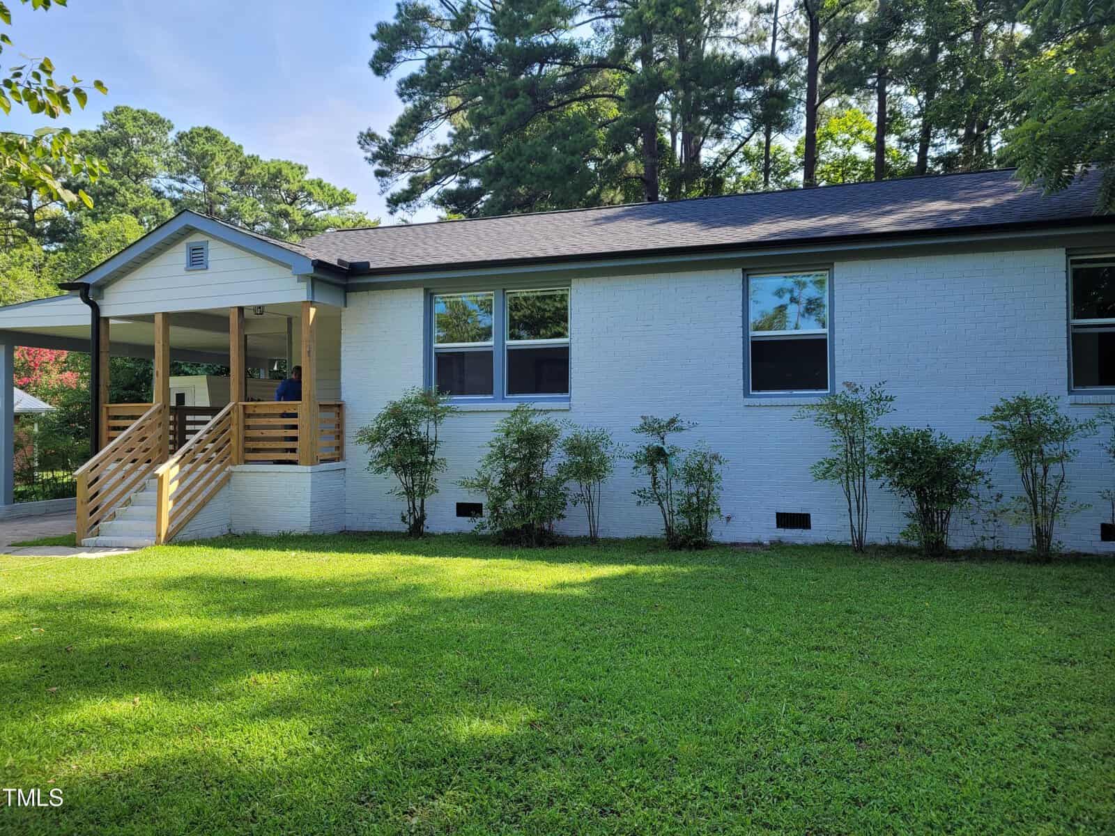 A white brick house at 308 9th Street, featuring a black shingled roof, is surrounded by green grass and shrubs. It has multiple windows and a covered porch with wooden railings. Tall trees rise in the background under a clear blue sky, embodying the charm of Butner.