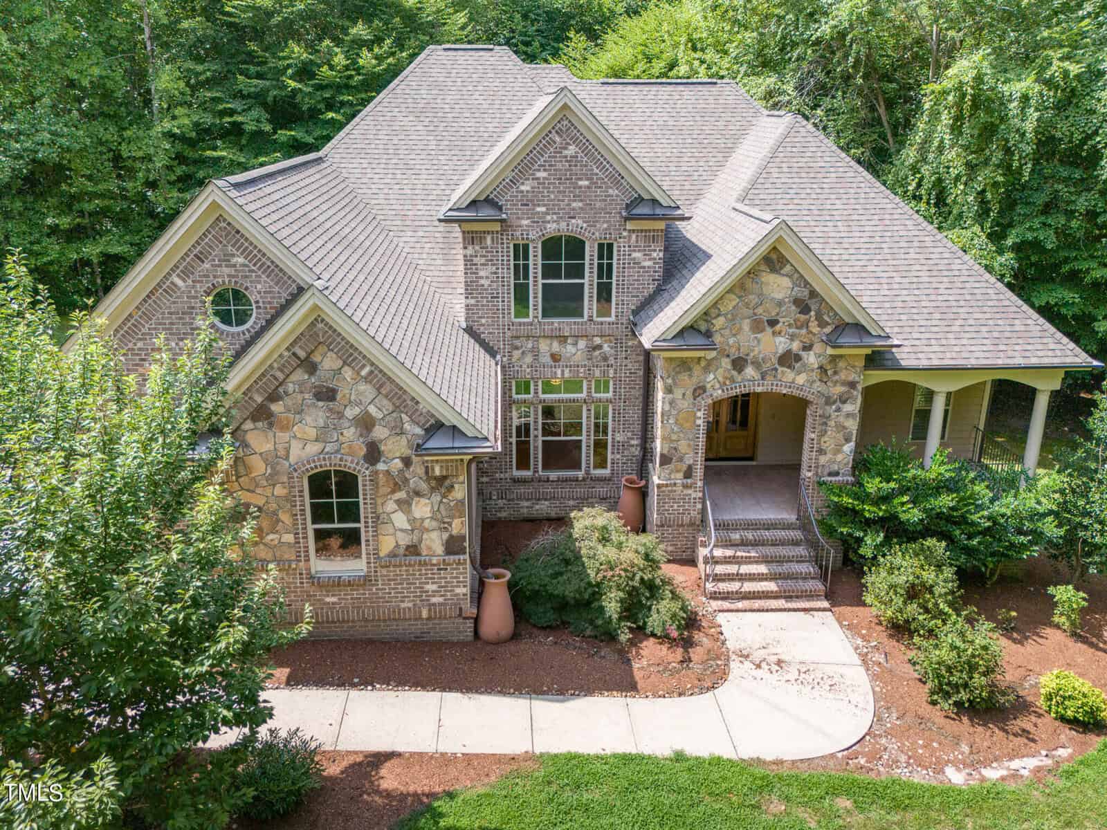A large, two-story brick and stone house with a gabled roof is surrounded by lush greenery and trees. A curved walkway leads to the front entrance, flanked by shrubs and potted plants. The house has multiple windows and detailed stonework.