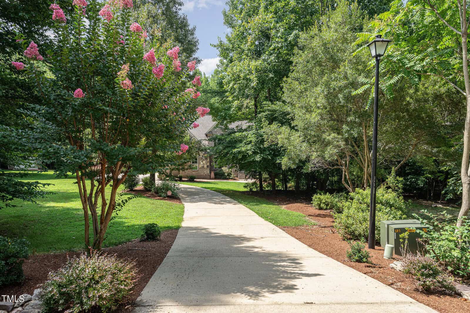 A paved pathway curves through a lush garden with green grass and various trees, including flowering ones. A lamp post is on the right, and a house is partially visible in the background under a clear blue sky.