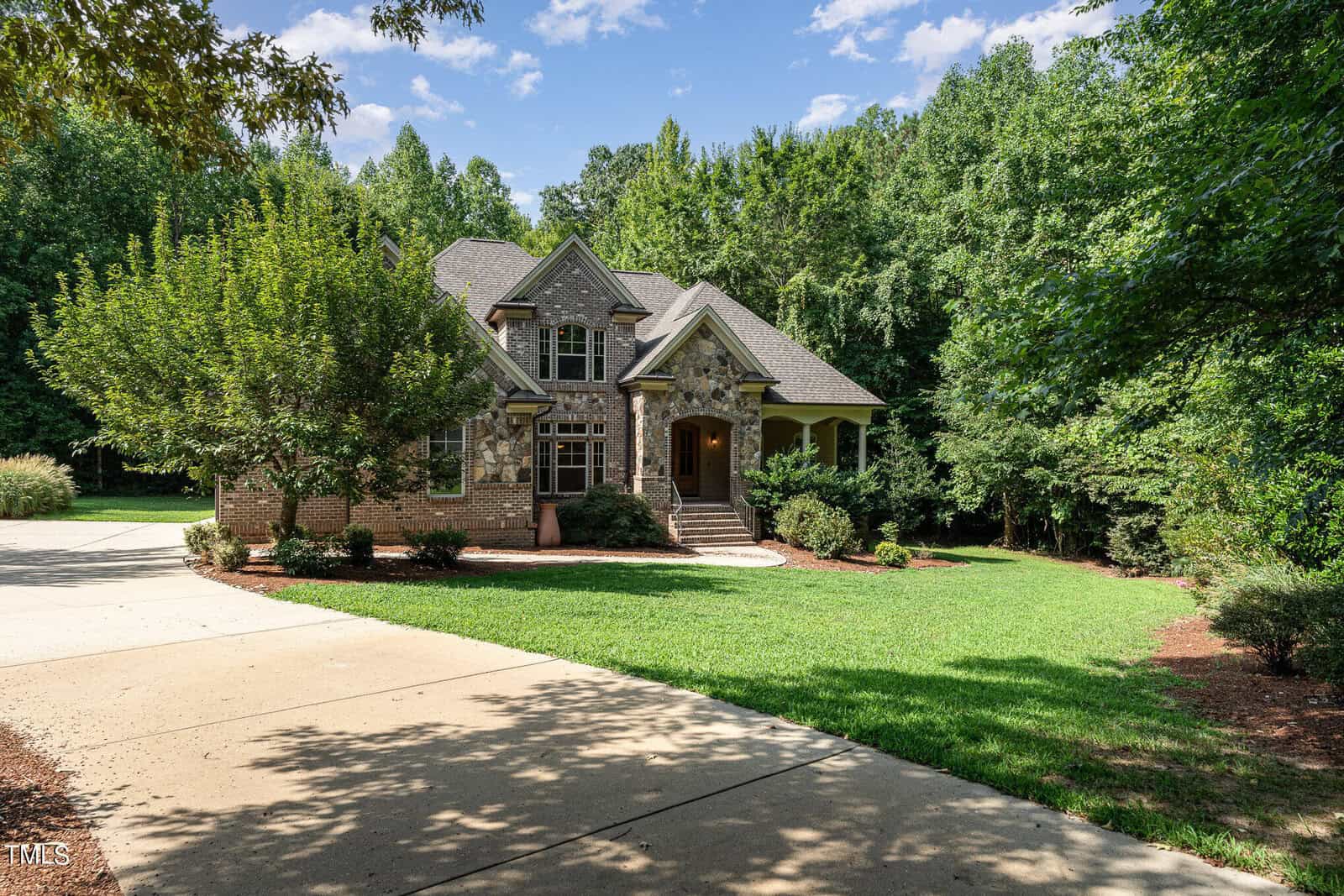 A large, stone and brick house with multiple gables is surrounded by lush greenery. The driveway leads up to a well-maintained front yard with trimmed bushes and trees under a partly cloudy blue sky.