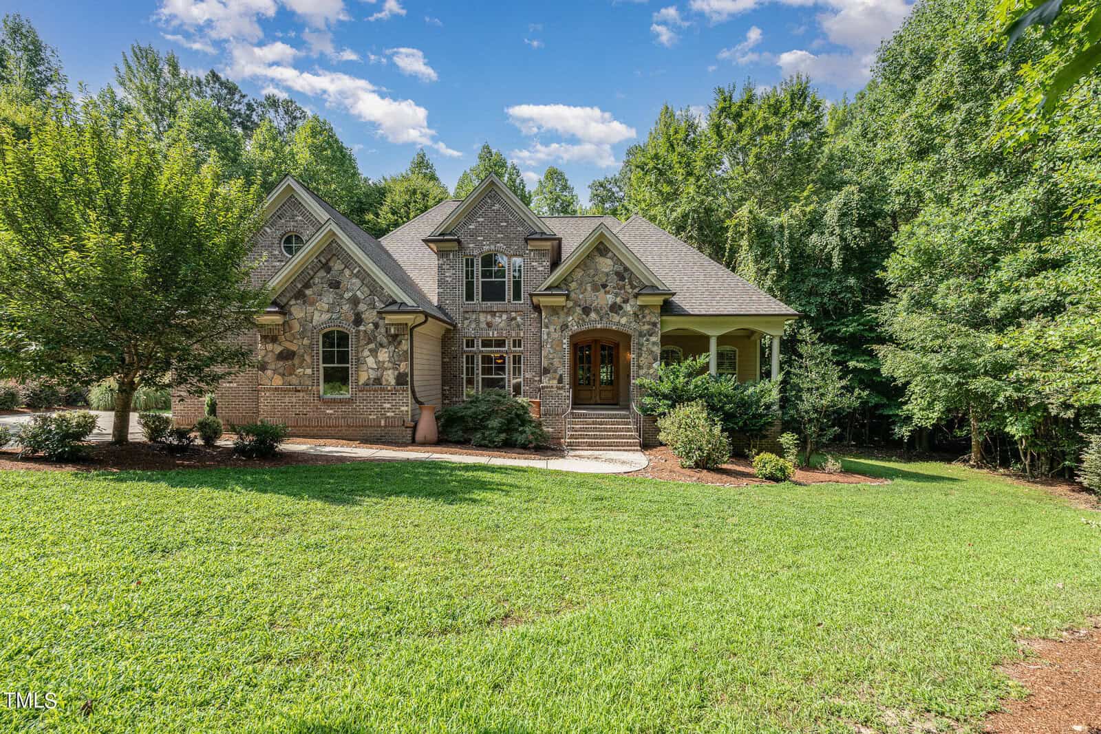A two-story brick and stone house at 1180 Oak Grove Court, Wake Forest, boasts arched windows and is enveloped by lush greenery. The front entrance features a small porch with steps leading to a wooden door, set against the backdrop of a clear sky with a few clouds.