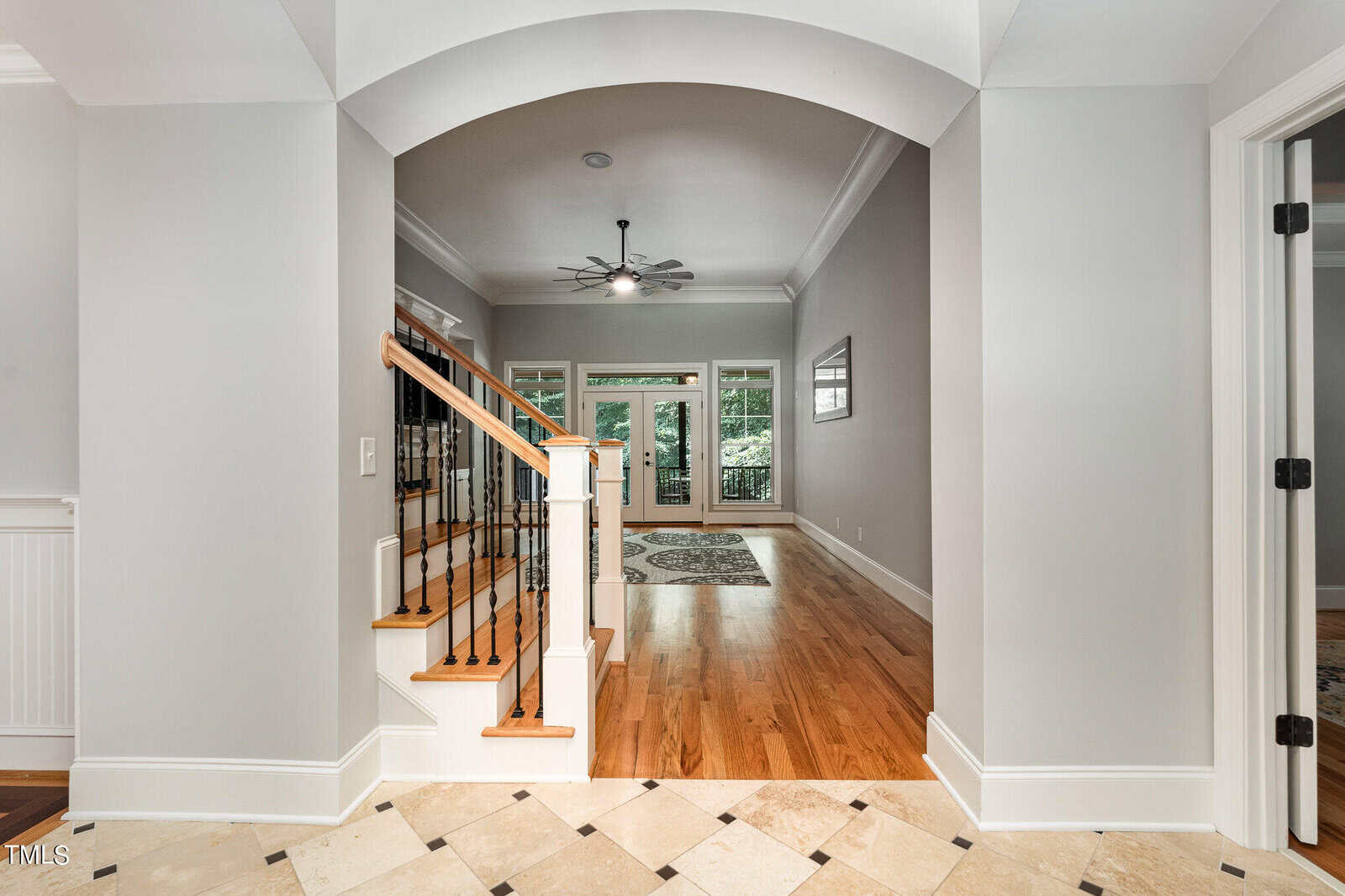 A hallway with a tiled floor leads to a room with wooden flooring. An archway frames a staircase on the left with wooden rails. Beyond, a ceiling fan hangs from the ceiling, and large windows show greenery outside. Doors are on the right.