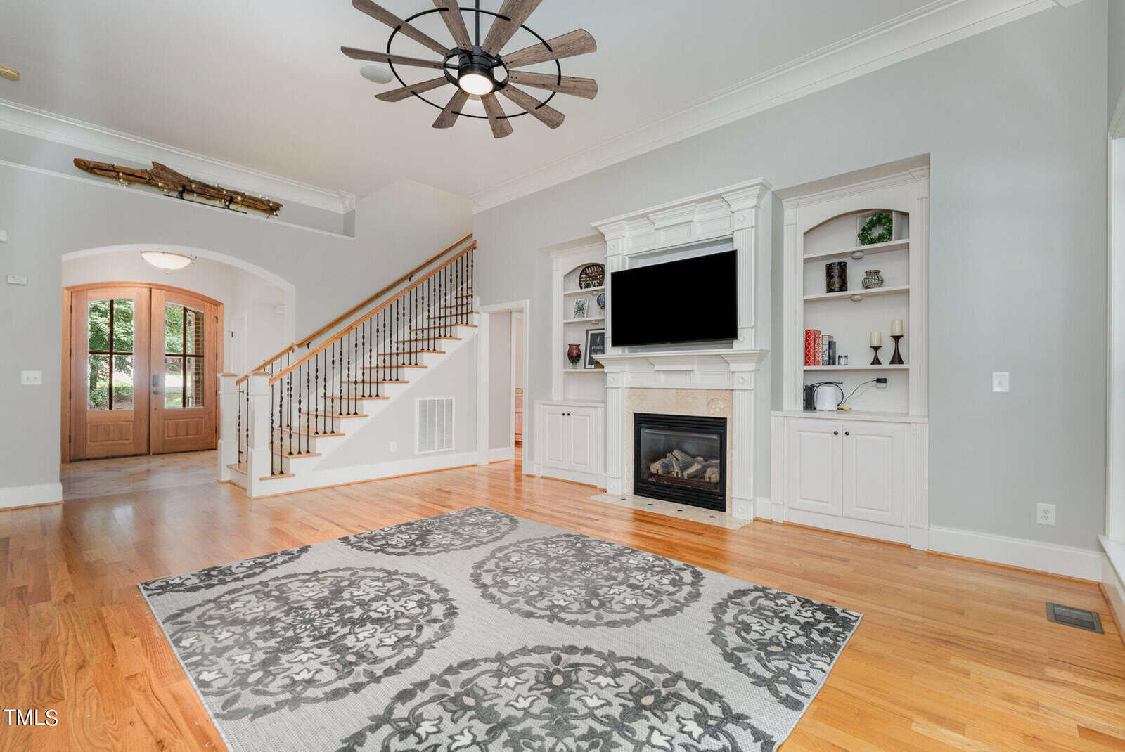 Spacious living room with hardwood floors, a large area rug, and a ceiling fan. Features a white fireplace with a TV above and built-in shelves on both sides. A staircase leads upstairs near a wooden front door with glass panels. Natural light fills the room.