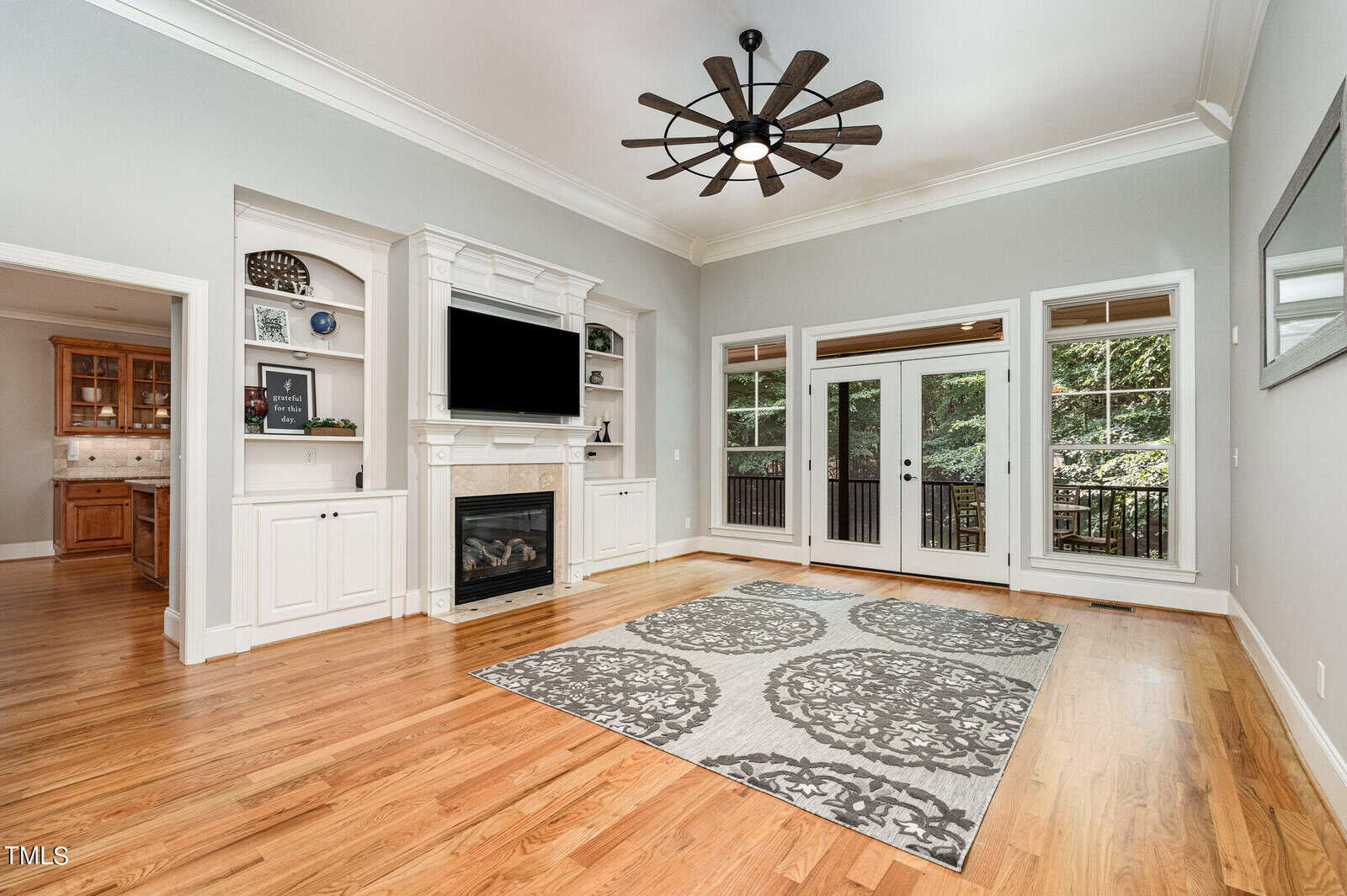 A spacious living room with hardwood floors, a large patterned rug, and a ceiling fan. The room features a fireplace with a TV above it, built-in shelves, and glass doors leading to a balcony with a view of trees. An open doorway leads to the kitchen.