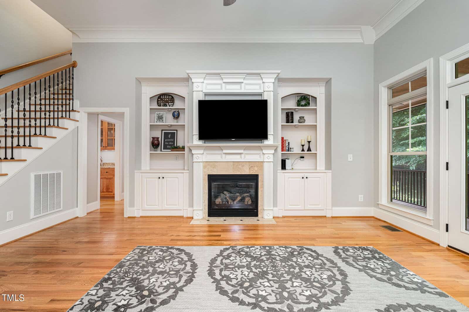 A spacious living room with a large, modern fireplace and a mounted TV. Built-in shelves surround the fireplace, displaying decor. A patterned rug covers the wooden floor. The space is bright with natural light from large windows. A staircase is on the left.