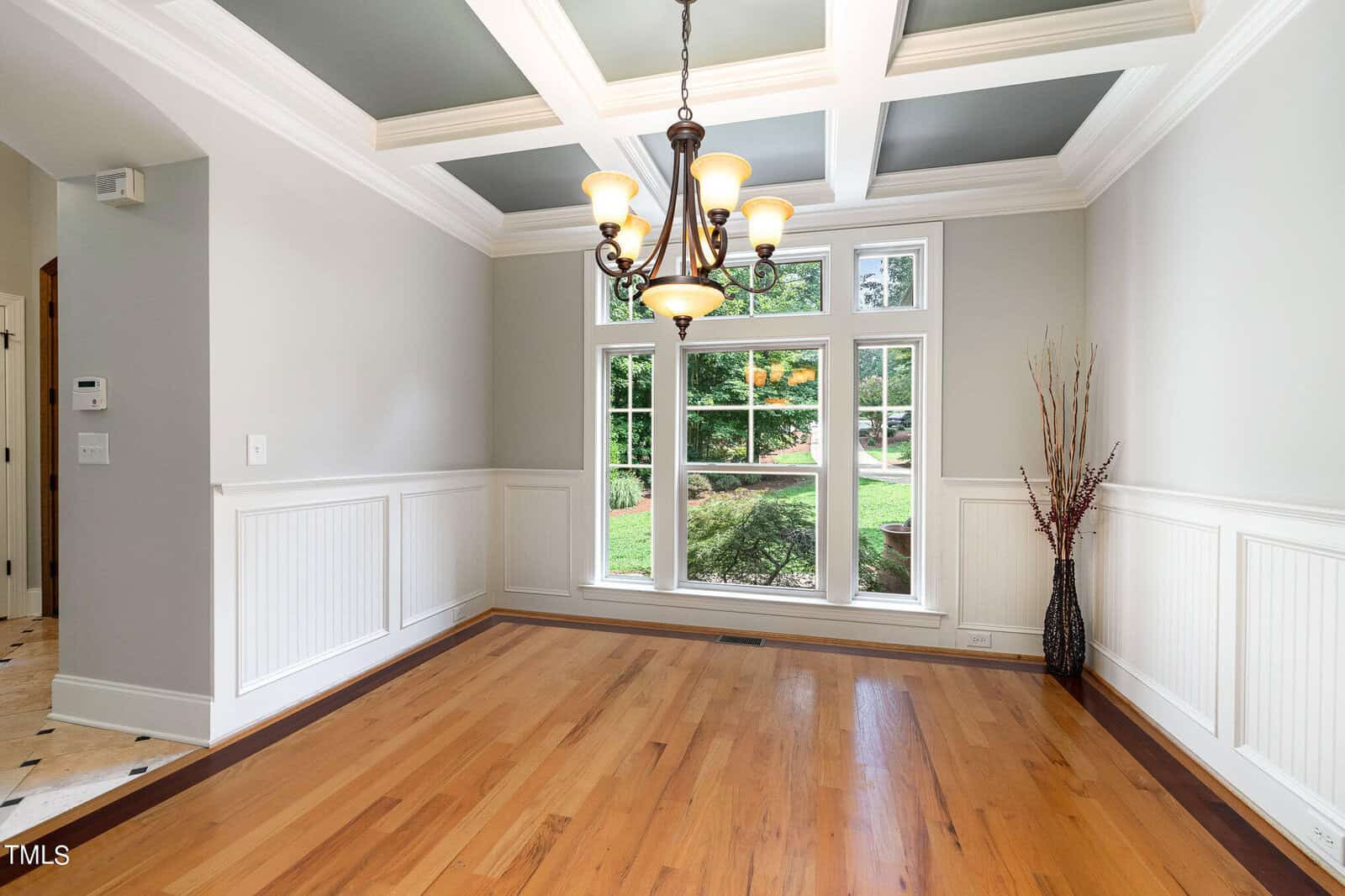 A bright dining room with a coffered ceiling and chandelier. The room has wainscoting, hardwood floors, and large windows offering a view of a garden. A decorative vase with branches stands in the corner.