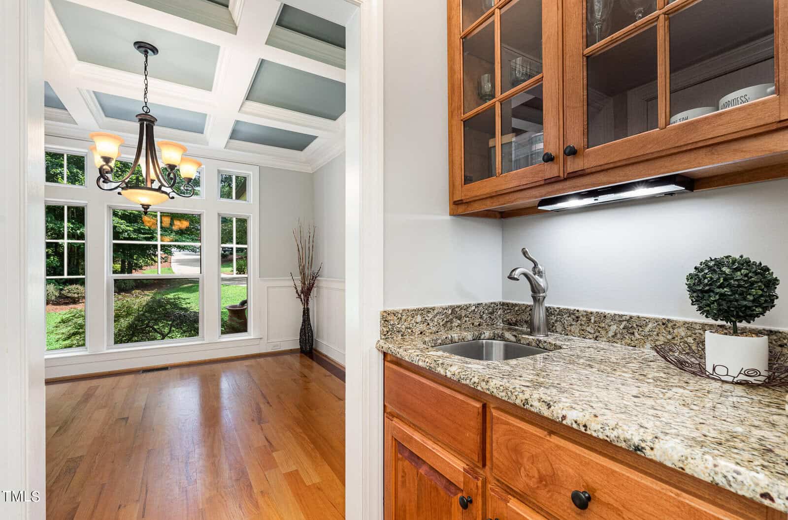 A kitchen area with granite countertops and wooden cabinets opens to a dining room with hardwood floors. A chandelier hangs from a coffered ceiling, large windows offer a view of greenery, and a small round plant decorates the counter.