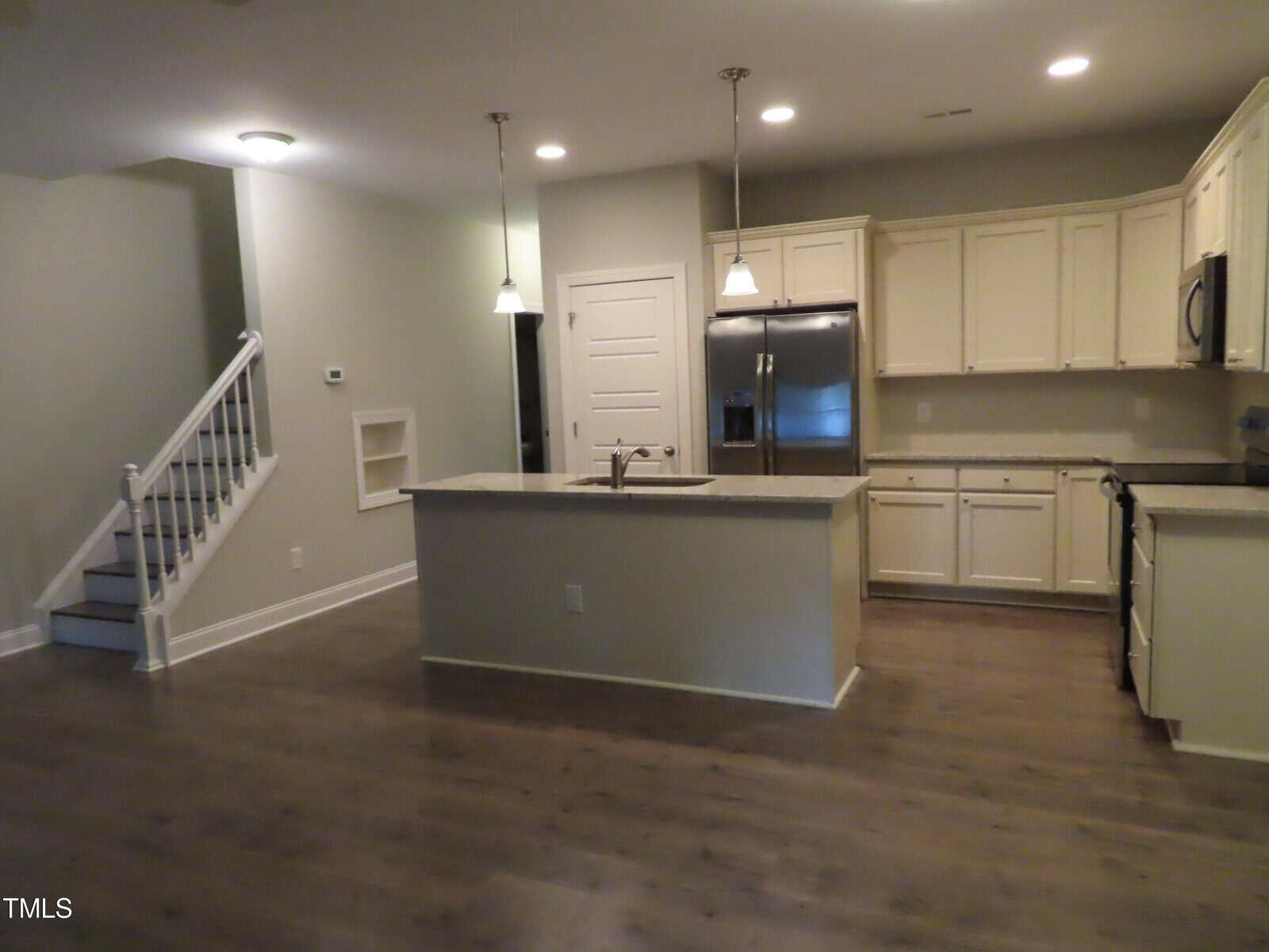 Modern kitchen with white cabinets and stainless steel appliances. A central island features a sink and pendant lighting. Dark hardwood floors contrast with light-colored walls. Stairs are visible to the left.
