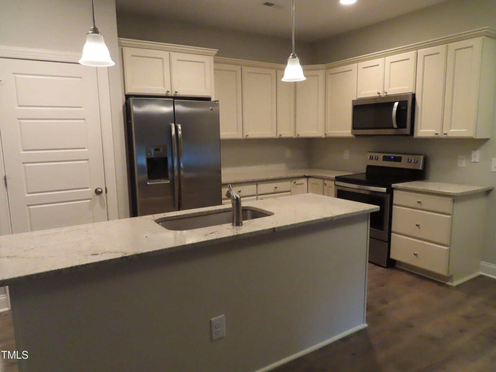 Modern kitchen with beige cabinets, stainless steel refrigerator, oven, and microwave. An island with a sink and pendant lighting hangs above a granite countertop. Wood flooring adds warmth to the space.