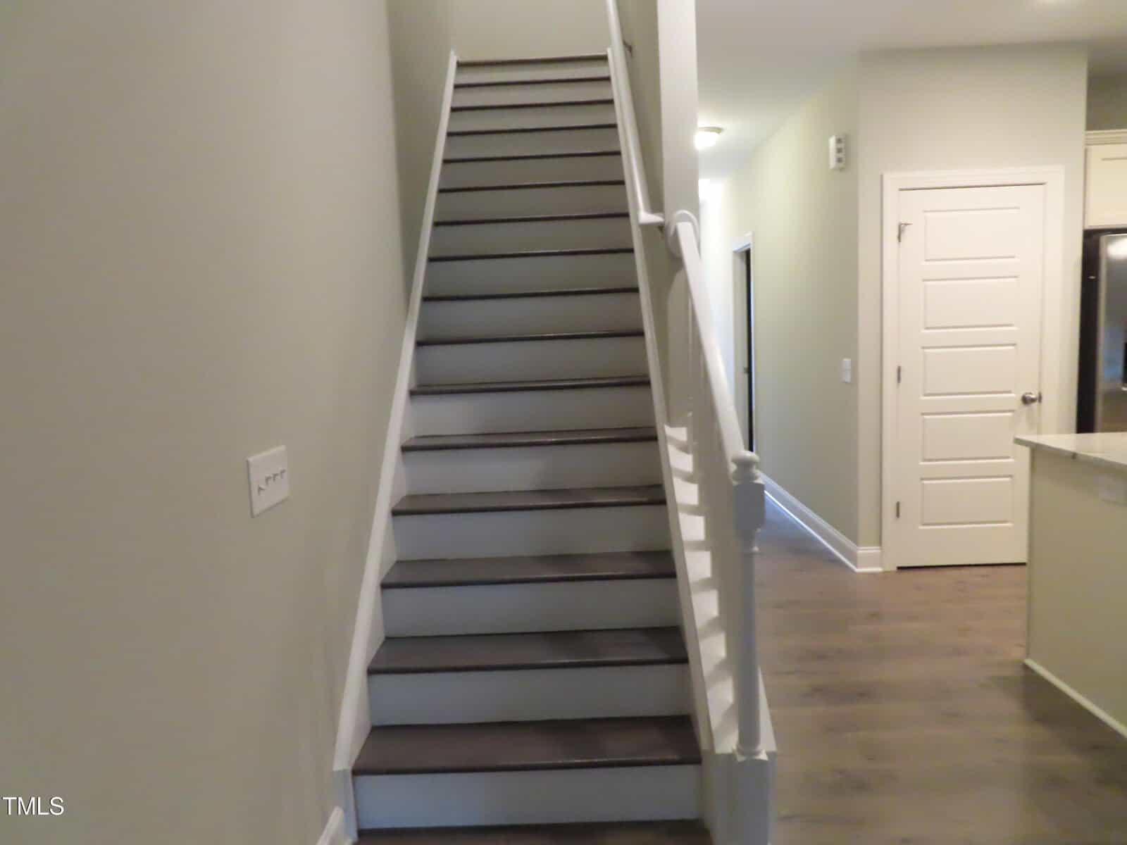 A modern interior staircase with wooden steps and a white handrail is next to a hallway. The walls are light-colored, and there is a closed door on the right side. The flooring is wood.