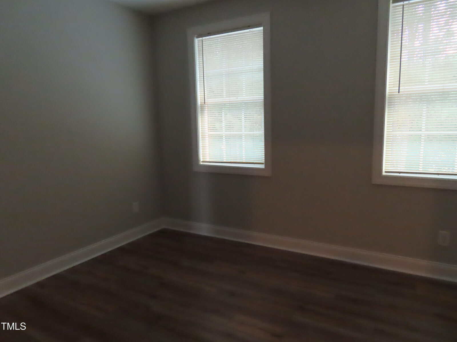 An empty room with two windows on adjacent walls, featuring closed blinds. The room has dark wooden flooring and light gray walls, creating a minimalist and clean look.