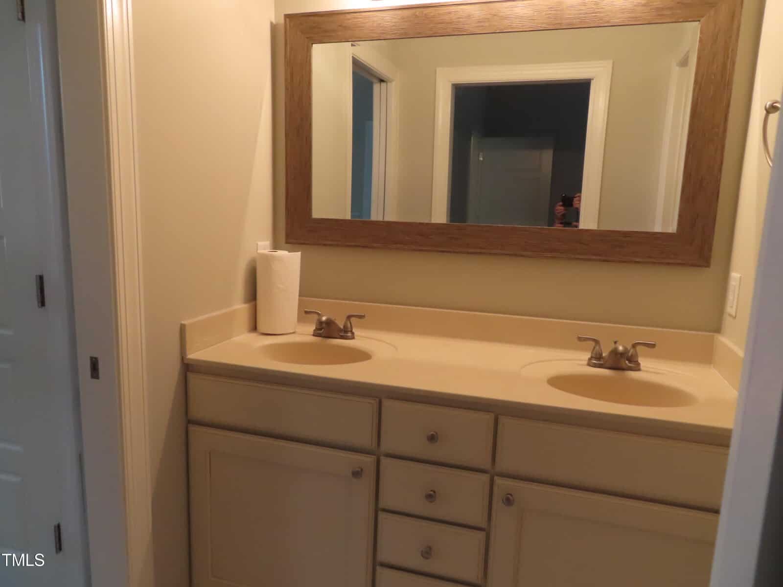 Bathroom with a double sink vanity. The countertop is beige, and there is a large mirror above it. A roll of paper towels sits on the left side of the counter. Walls and cabinets are a light color.