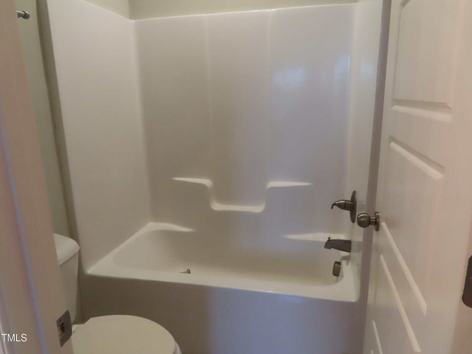 A small bathroom featuring a white bathtub with a shower. The walls are light-colored, and the bathroom door is partially open. A toilet is visible in the foreground.