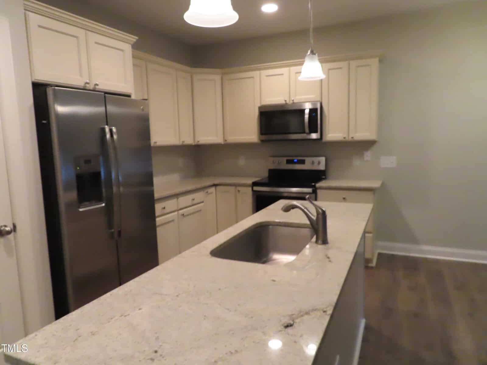 Modern kitchen with a gray granite island featuring a sink and faucet. Stainless steel appliances, including a double-door fridge and oven with a microwave above, are set against beige cabinetry. A pendant light hangs above the island.