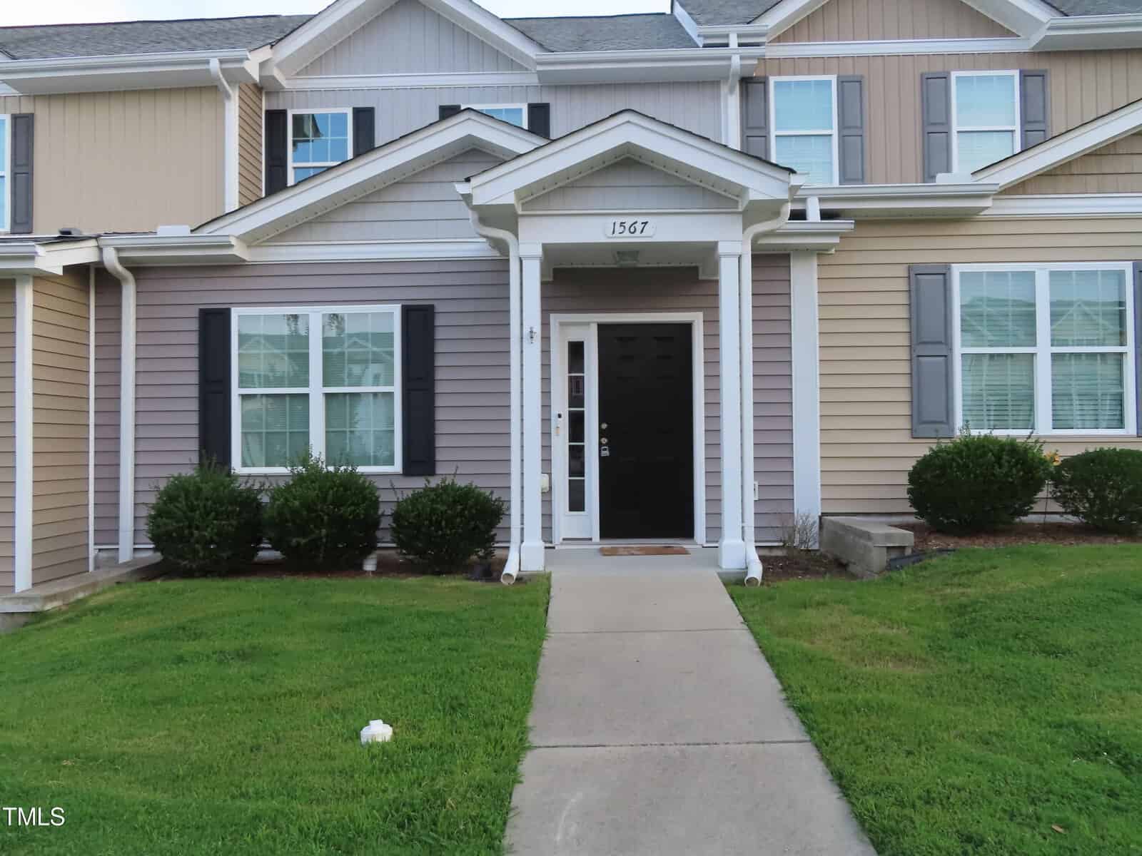 A charming two-story townhouse at 1567 Quail Circle boasts beige and cream siding. The small porch features a black front door and white columns. Lush green bushes adorn the front garden, while a concrete path guides you to the entrance.