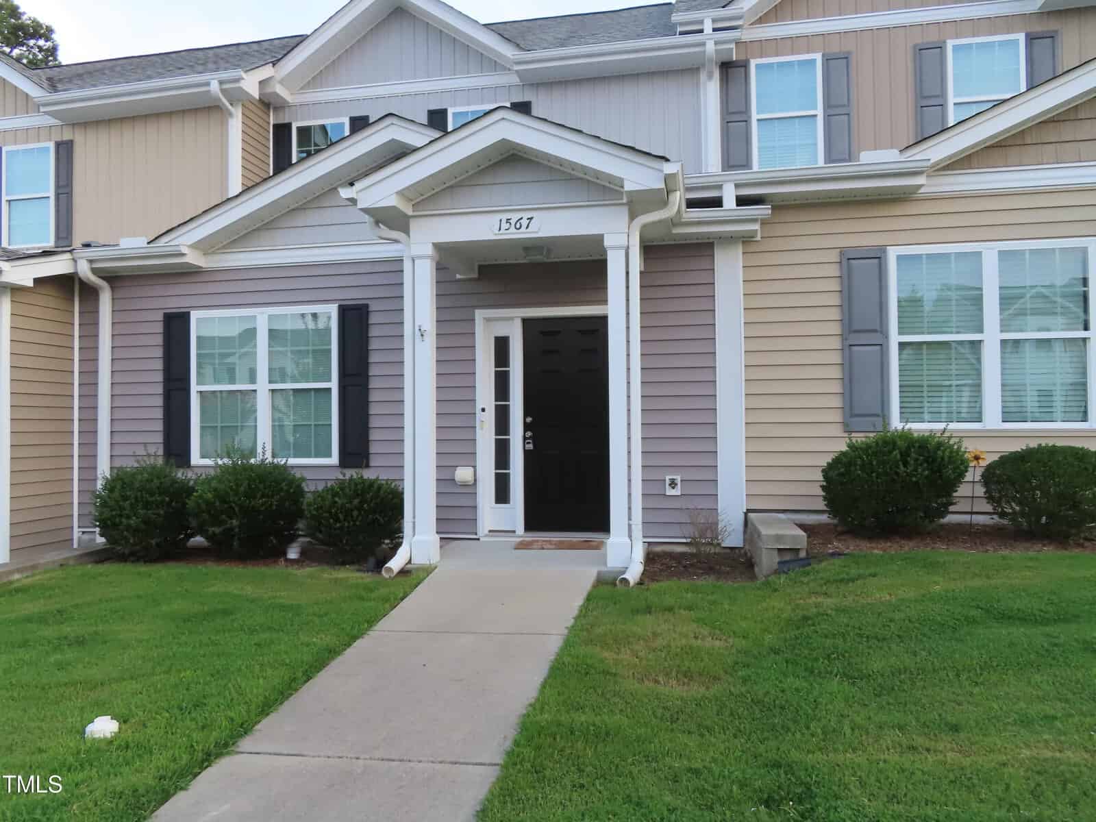 Image of a two-story townhouse with tan and beige siding. The front features a black door with white trim, small porch, and symmetrical windows with black shutters. Green bushes and a well-kept lawn line the walkway leading to the entrance.