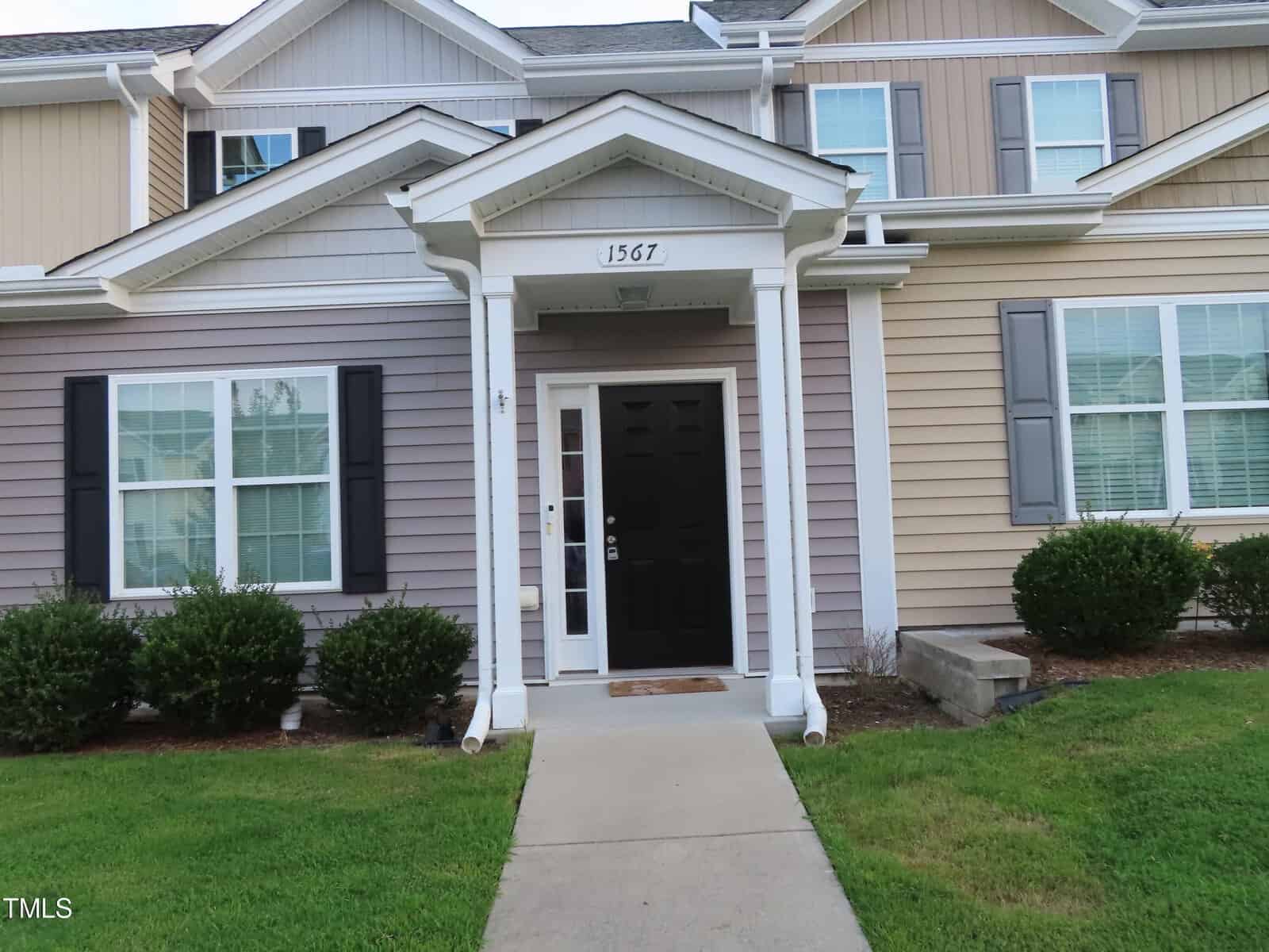 A two-story townhouse with beige and gray siding, black front door, and white trim. The entrance is framed by two columns and a small roof overhang. The house number "1567" is displayed above the door. Bushes line the walkway leading to the entrance.